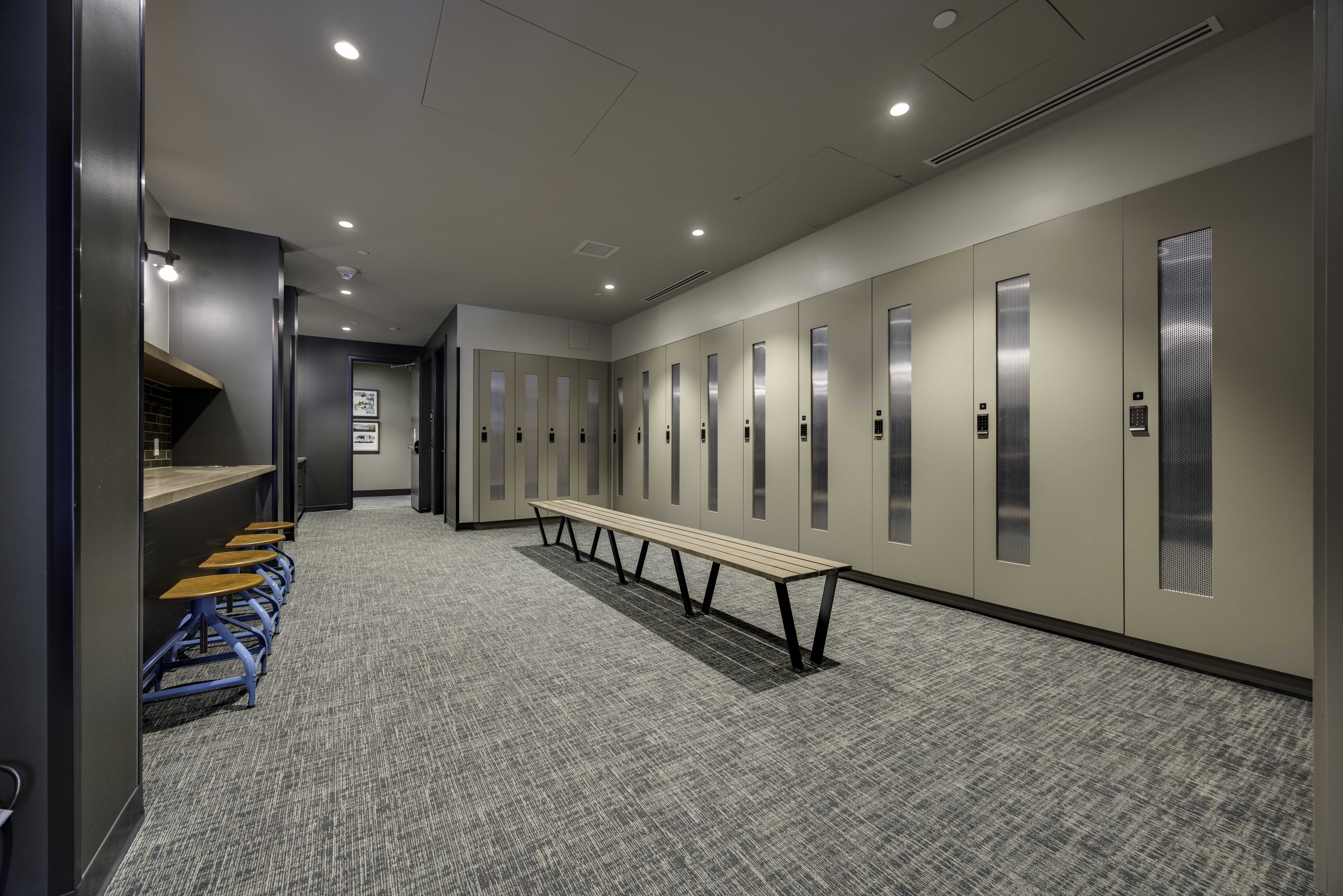 This modern locker room facility features a long, central wooden bench with a black metal frame, flanked by a wall of tall, neutral-toned lockers with integrated digital locks and vertical glass panels. To the left, a counter area with blue-framed stools provides a convenient space for seating or personal items, while the room is finished with a textured grey carpet. The perspective is a wide-angle shot looking down the length of the room, emphasizing the clean, organized, and professional aesthetic of the amenity space.
