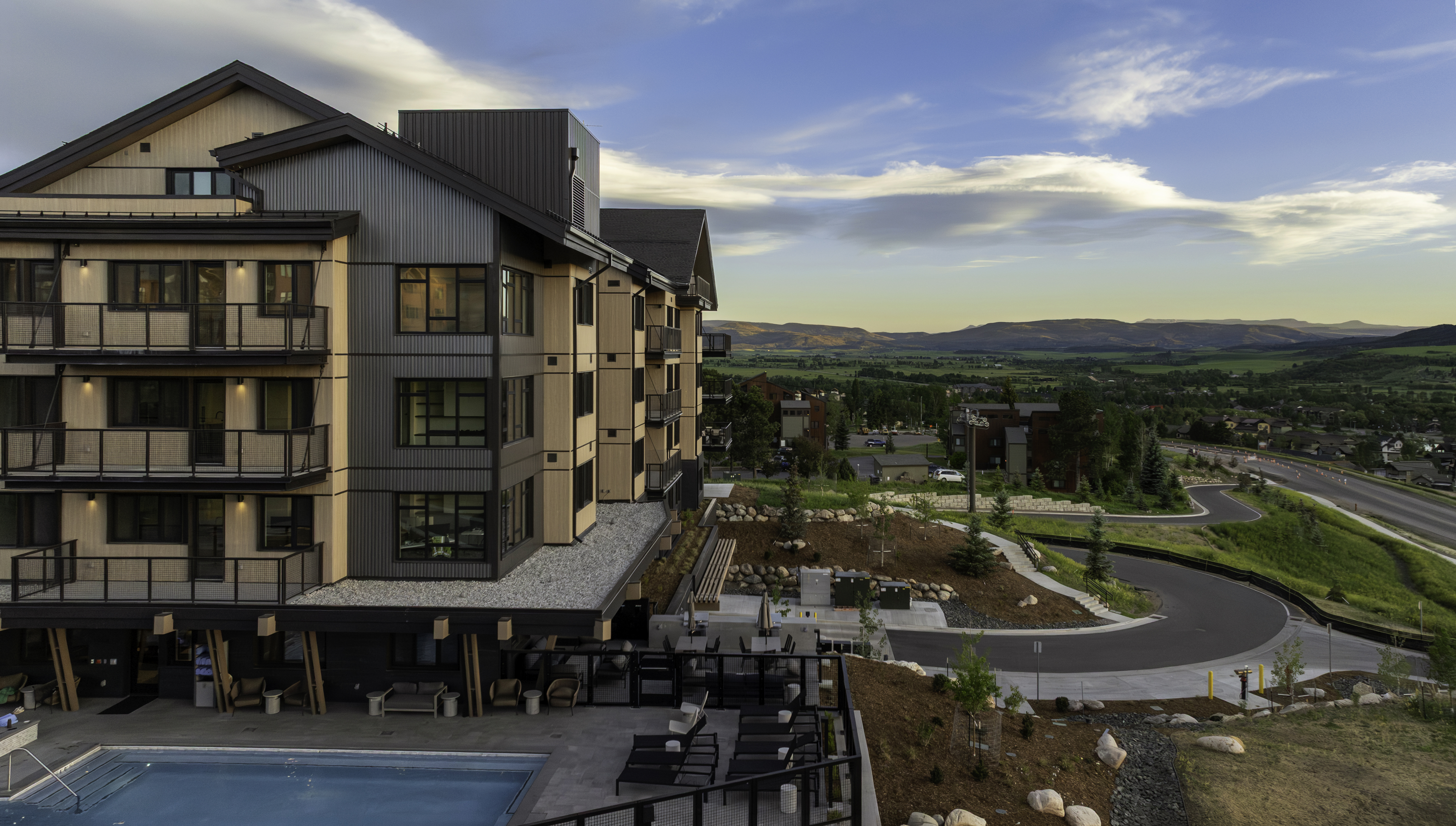 This elevated perspective captures a modern multi-story residential building featuring a mix of light wood-toned siding and dark corrugated metal panels. The structure includes stacked balconies and a ground-level patio area with a swimming pool, all set against a scenic backdrop of rolling hills and a valley at sunset. A winding road curves through the landscaped grounds, emphasizing the property's integration into the surrounding natural landscape.