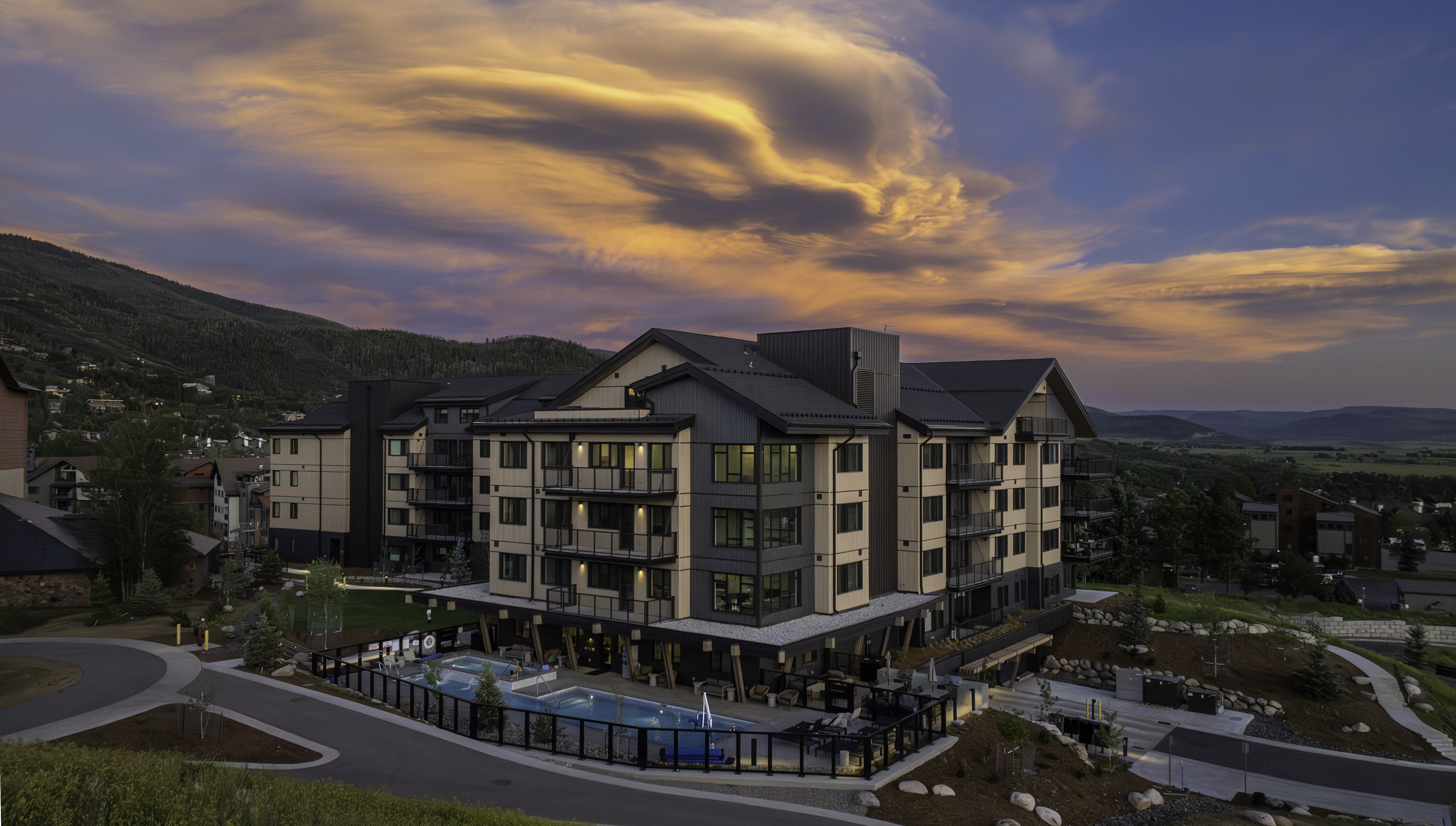This elevated aerial perspective captures a modern multi-story residential complex set against a dramatic sunset sky. The building features a contemporary design with a mix of light-toned siding, dark metal accents, and numerous balconies, while a pool and spa area are situated at the base. The surrounding landscape includes paved driveways, rocky terrain, and a distant mountain backdrop, creating a serene and upscale atmosphere.