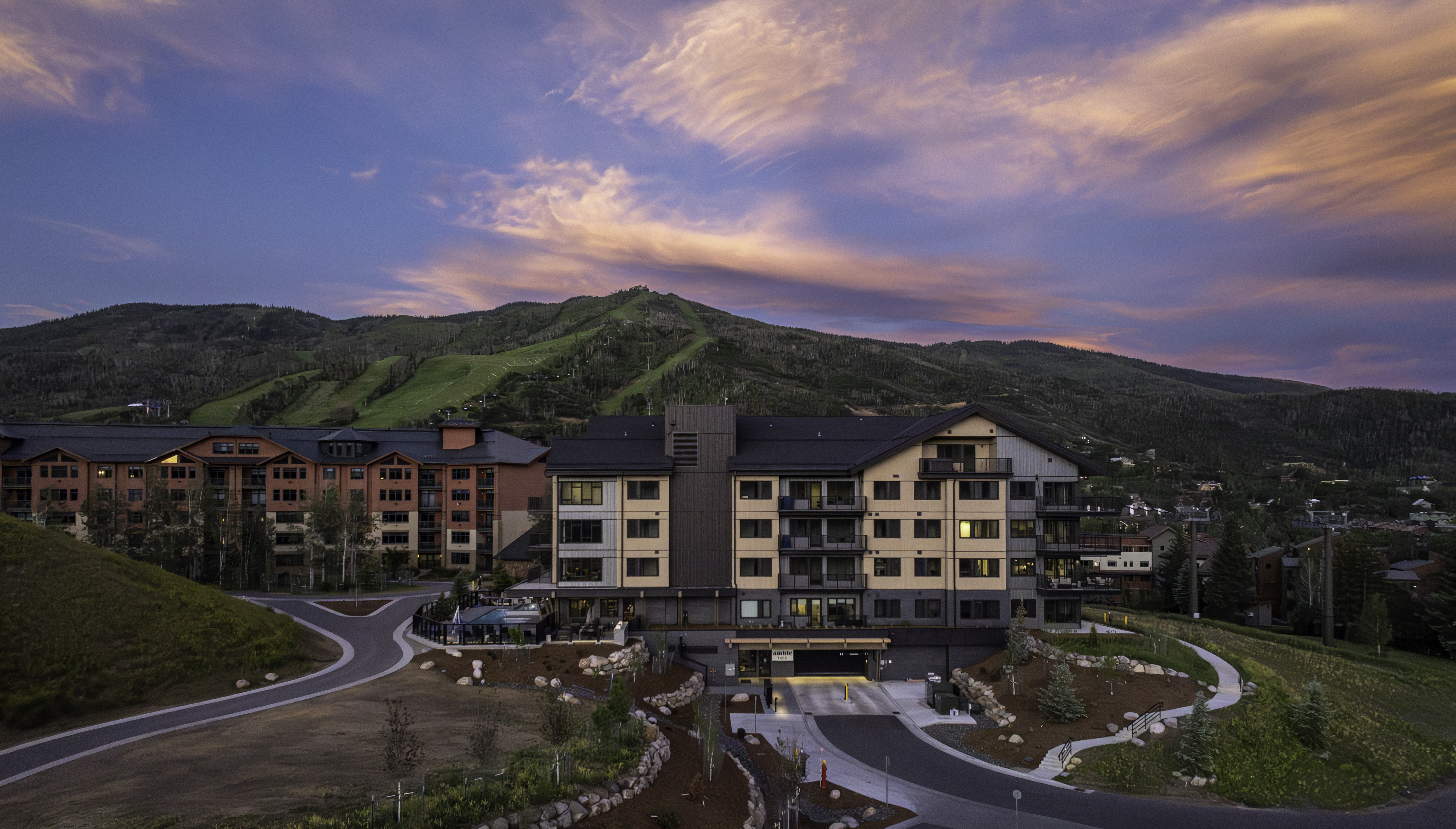 This aerial view captures a modern multi-story residential complex nestled at the base of a lush, green mountain range during a vibrant sunset. The architecture features a blend of contemporary materials, including dark metal accents and light-toned siding, with multiple balconies and a ground-level parking entrance. A small outdoor pool area is visible to the left, and the surrounding landscape includes paved driveways and natural, terraced rock landscaping.