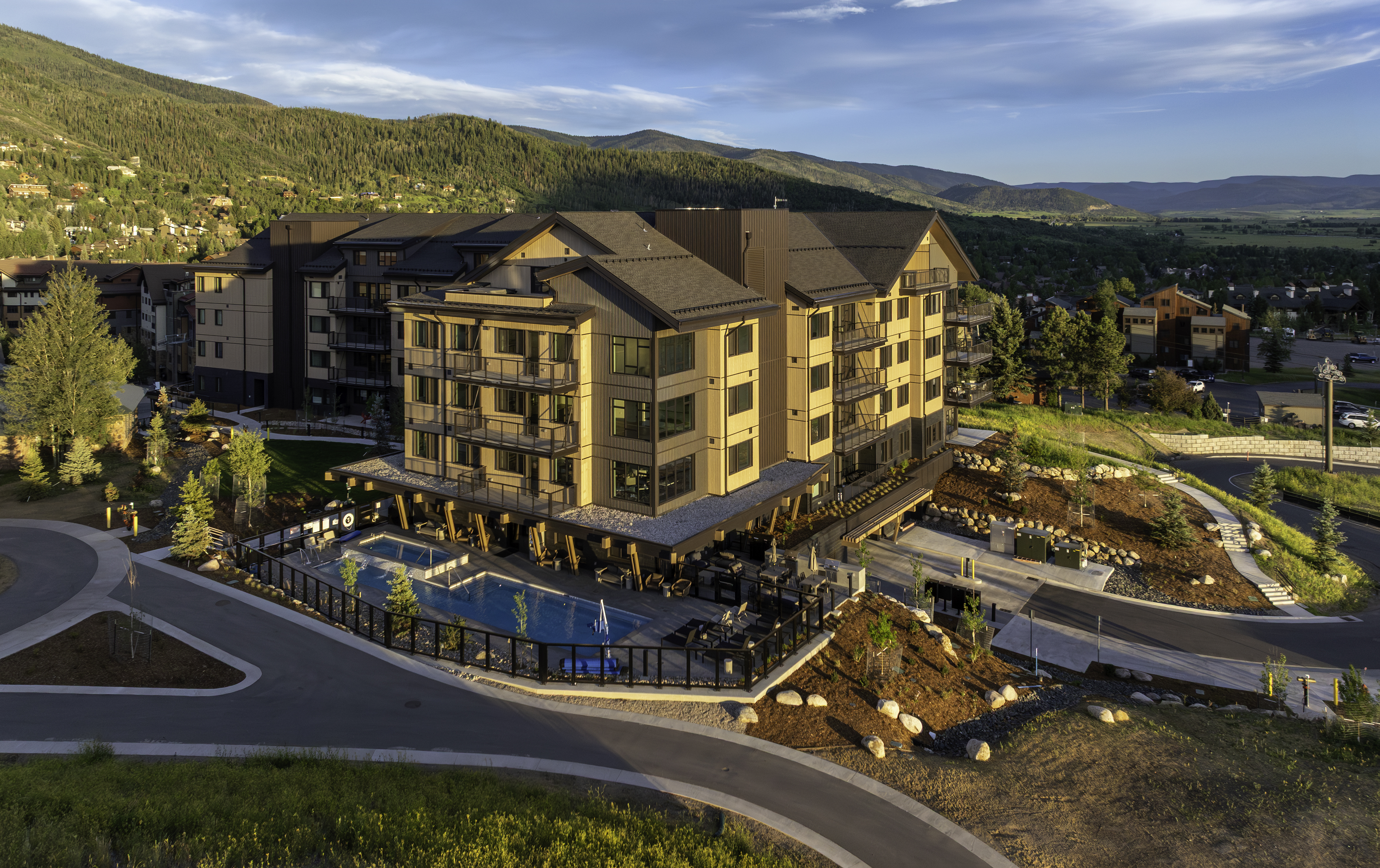 This high-angle aerial view captures a modern, multi-story mountain resort building nestled against a backdrop of rolling, forested hills. The structure features a contemporary design with a mix of wood-toned siding and dark metal accents, complemented by a swimming pool and spa area on the lower level. The surrounding landscape includes paved pathways, native vegetation, and a clear view of the valley, creating an inviting and serene atmosphere.