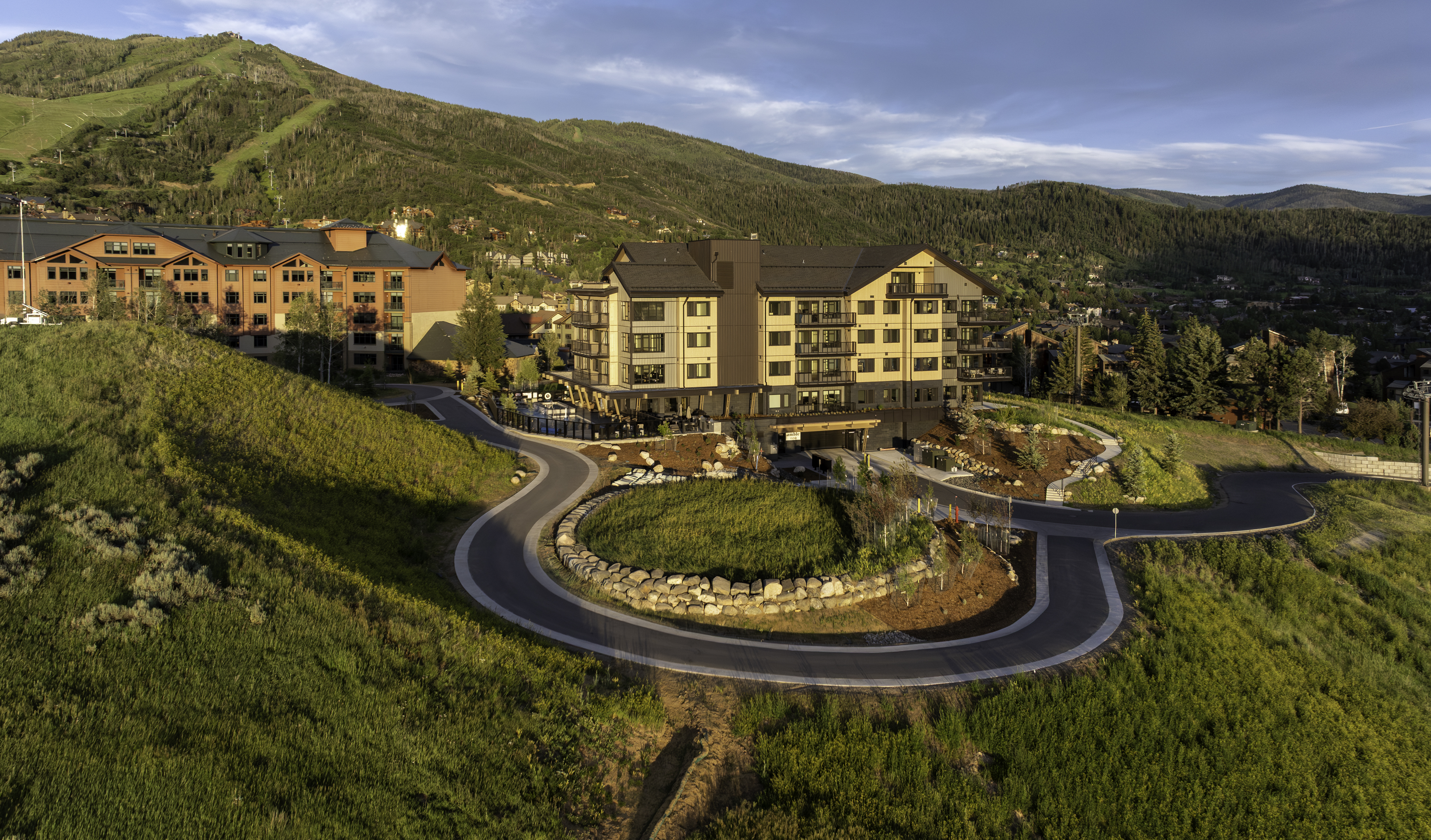 This high-angle aerial shot captures a modern multi-story resort complex nestled into a lush, mountainous landscape during the golden hour. The architecture features a blend of light-toned siding and dark accents, with a winding paved driveway leading to the main entrance and a lower-level parking area. The surrounding terrain is characterized by rolling green hills and dense forest, creating a serene and upscale mountain retreat atmosphere.