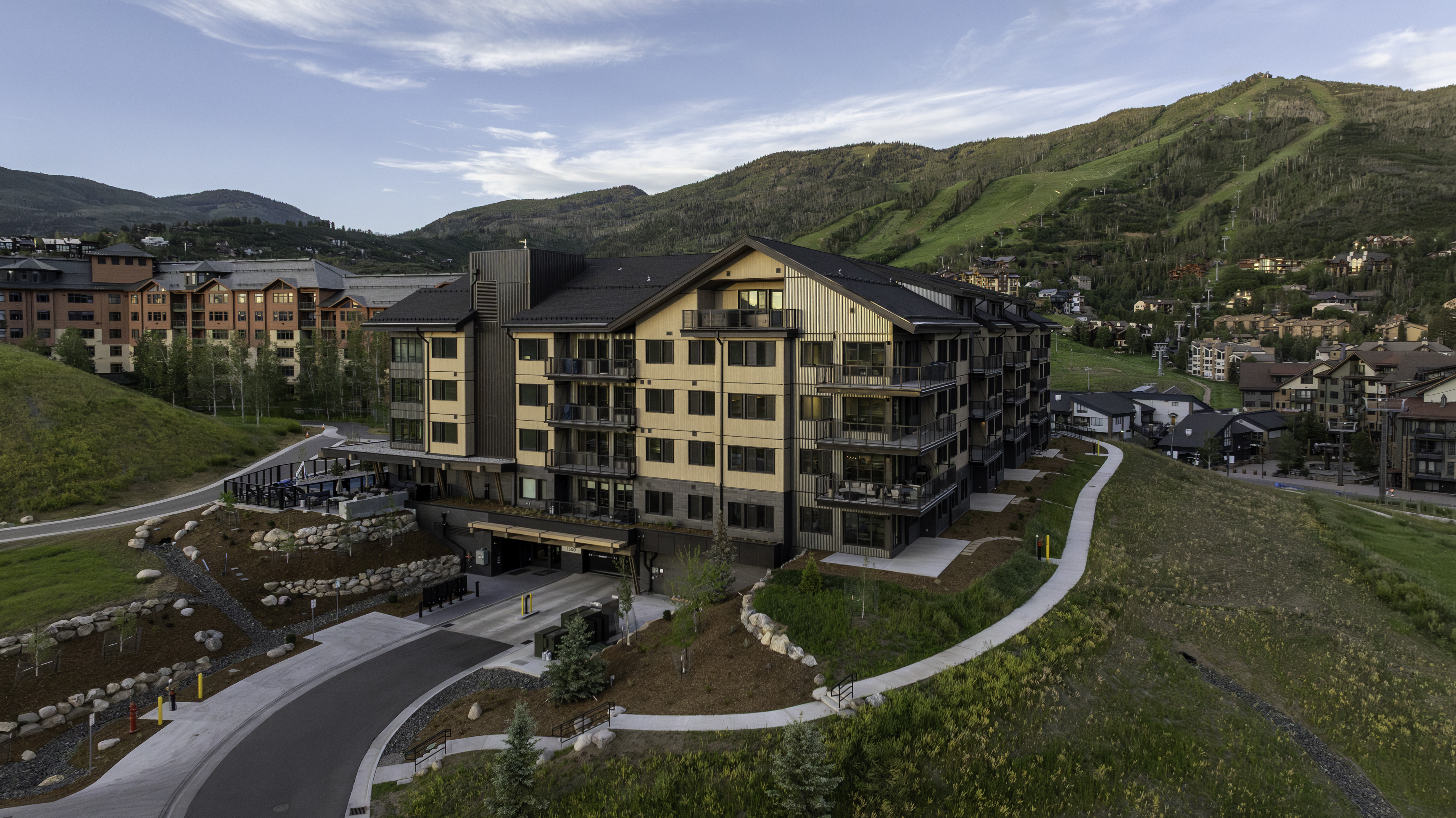 This high-angle aerial view captures a modern, multi-story mountain resort complex nestled against a lush, green ski slope. The building features a contemporary design with a mix of dark metal siding, light-toned panels, and numerous private balconies, complemented by a paved driveway and a landscaped entrance. The surrounding landscape includes rolling hills, scattered trees, and other residential properties, creating a serene and upscale alpine atmosphere.