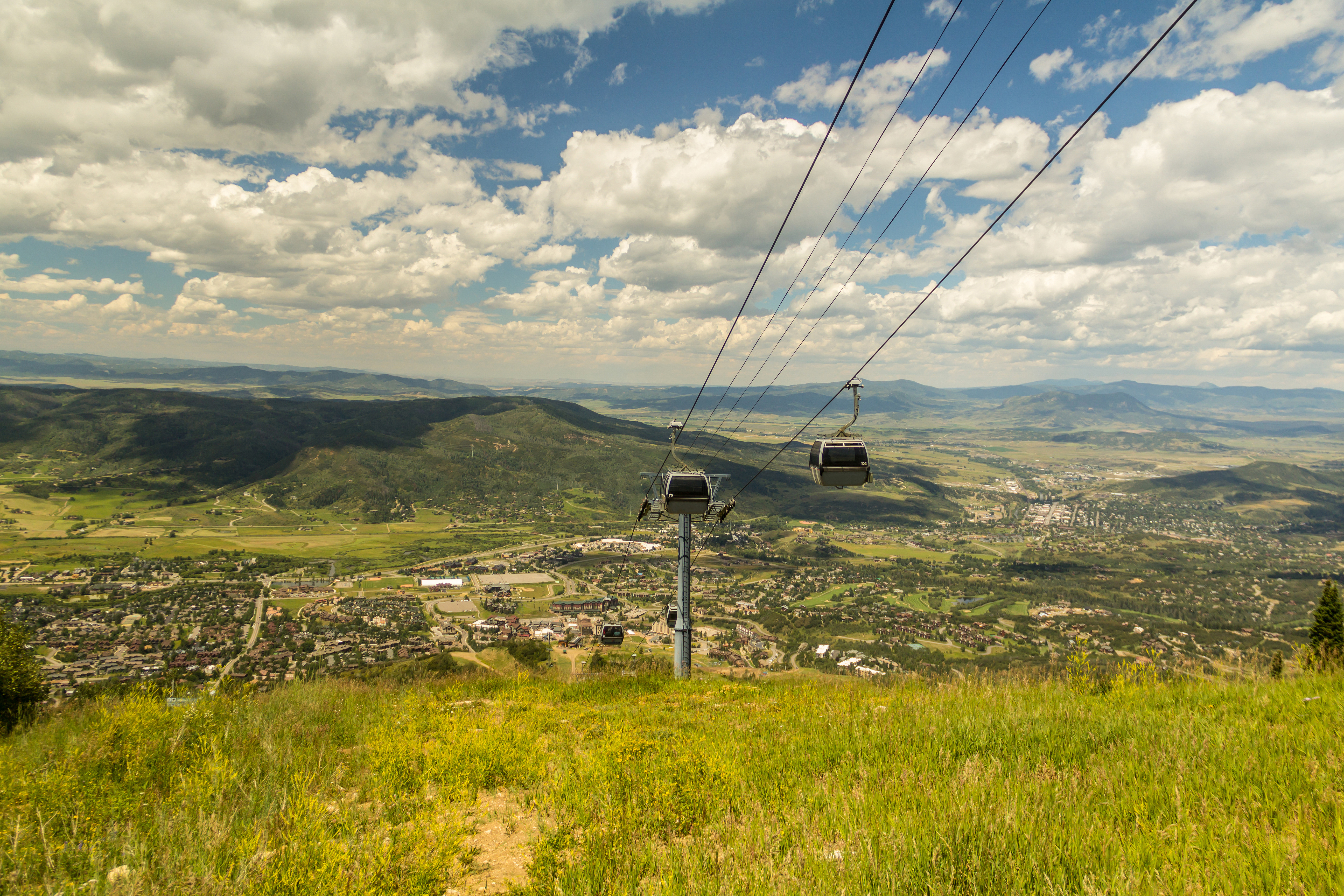 This high-altitude, wide-angle shot captures a sweeping panoramic view of a mountain valley town from a grassy hillside vantage point. In the foreground, a gondola lift system with suspended cabins cuts through the frame, leading the eye toward the sprawling residential community and lush green landscape below. The scene is bathed in bright daylight under a vast, cloud-dappled sky, emphasizing the expansive scale and scenic location of the property.