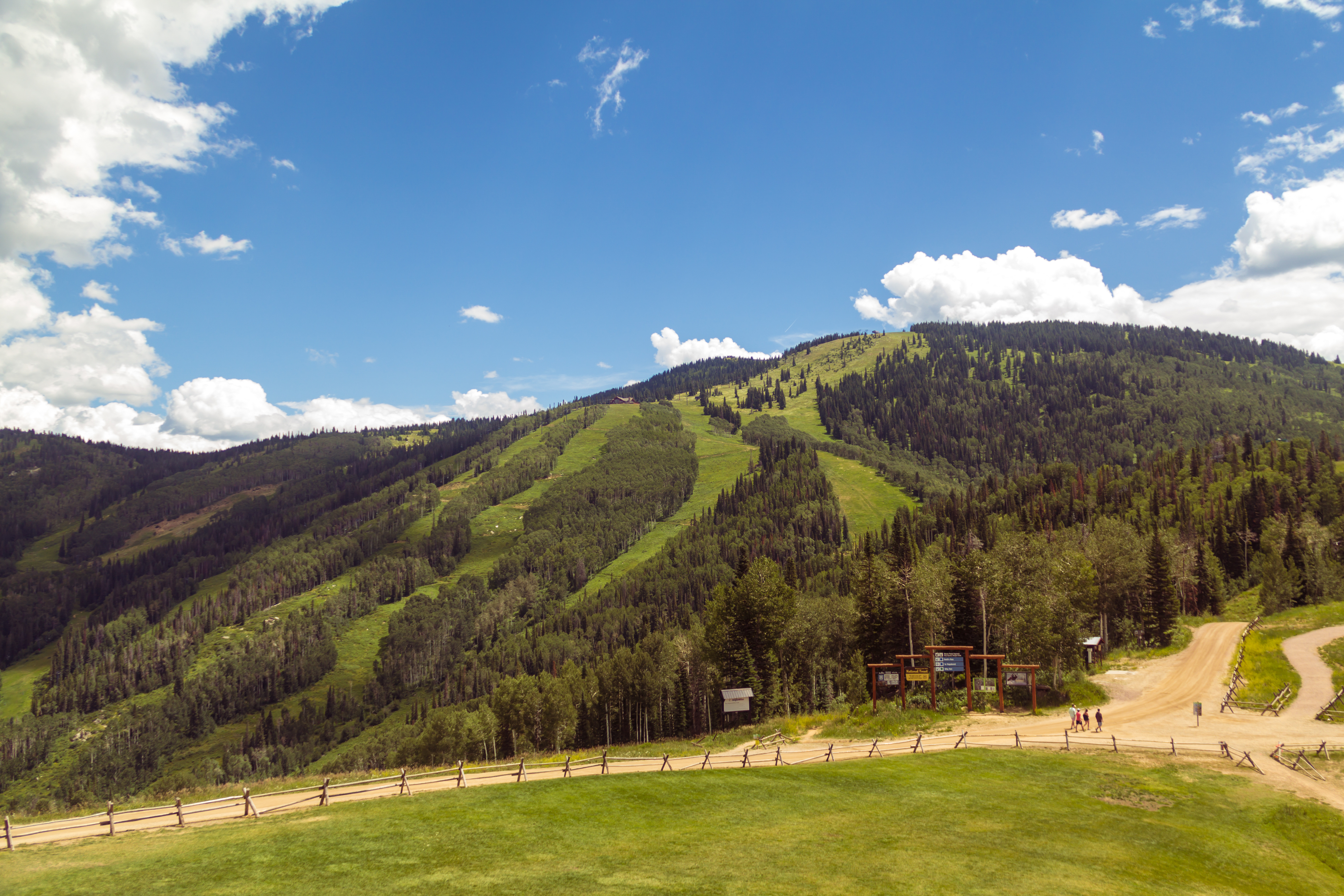 This wide-angle shot captures a scenic mountain landscape featuring a ski resort slope covered in lush green vegetation and dense pine forests. In the foreground, a manicured grassy lawn is bordered by a rustic wooden fence, leading toward a dirt trail where a small group of people is walking near a trailhead sign. The perspective is elevated, providing a sweeping, cinematic view of the expansive terrain under a bright blue sky with scattered white clouds.