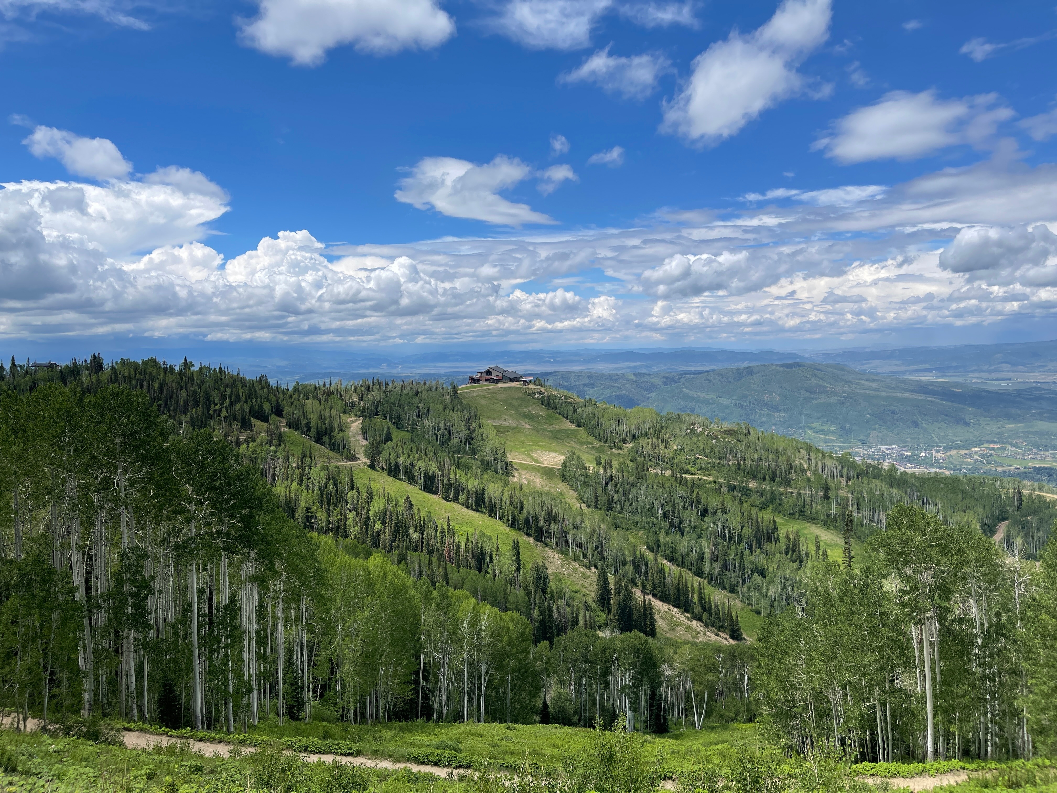This wide-angle landscape shot captures a mountain resort setting, featuring a prominent lodge perched atop a lush, green ridge. The foreground is dominated by a dense forest of aspen and pine trees, while the mid-ground reveals ski runs carved into the hillside leading up to the structure. The expansive blue sky filled with dramatic, fluffy white clouds provides a scenic backdrop for this elevated, panoramic view.