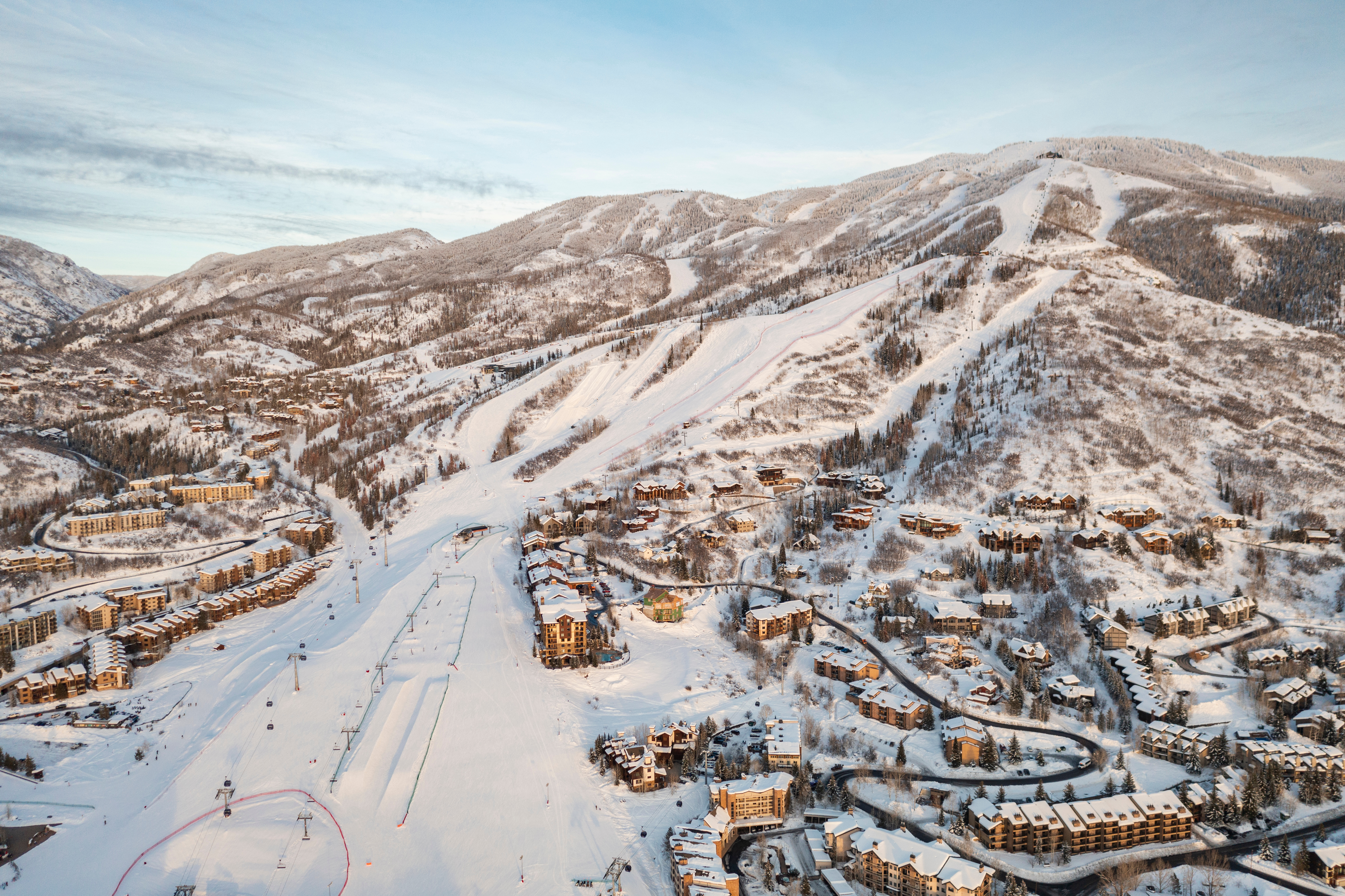 This expansive aerial view captures a sprawling mountain resort nestled within a snow-covered landscape. The scene features numerous multi-story residential buildings and lodges integrated into the hillside, alongside groomed ski runs and chairlift infrastructure. The golden hour light casts long shadows across the terrain, highlighting the rugged topography and the dense evergreen forests surrounding the development.