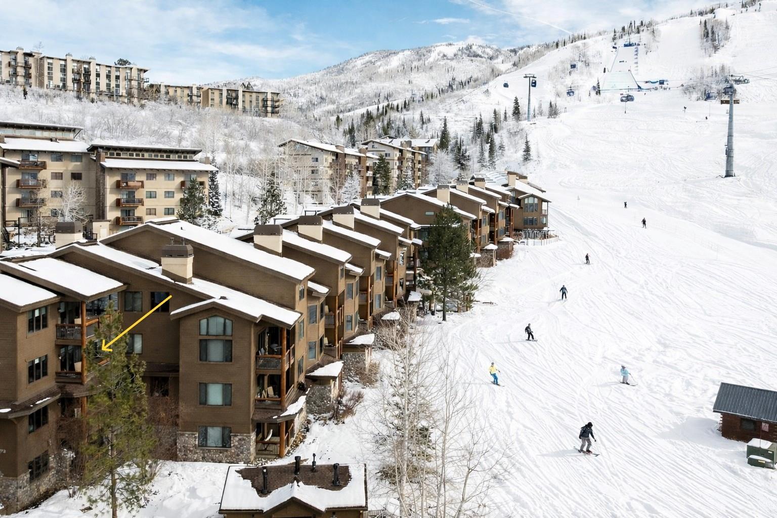 This high-angle aerial shot captures a multi-story mountain resort complex nestled directly against a snowy ski slope. The architecture features rustic wood siding, stone foundations, and multiple balconies, with a yellow arrow highlighting a specific unit's outdoor space. Skiers are visible navigating the groomed run, emphasizing the property's prime ski-in/ski-out location and scenic alpine setting.