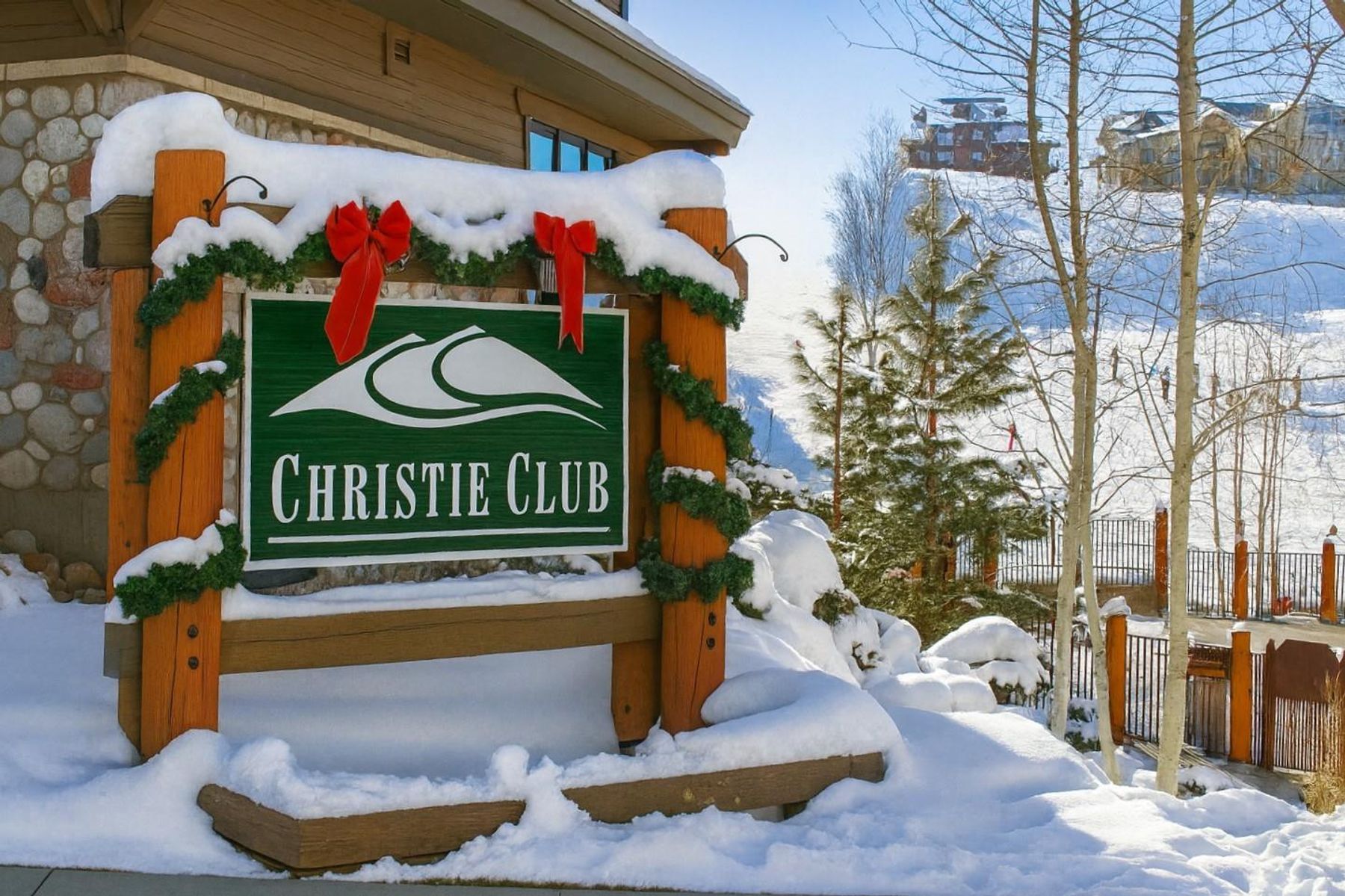This image captures the entrance sign for the Christie Club, a mountain resort property set against a backdrop of snow-covered slopes and evergreen trees. The wooden sign is decorated with festive greenery and red bows, emphasizing a welcoming, seasonal atmosphere. The perspective is a ground-level shot looking toward the resort grounds, highlighting the snowy landscape and nearby residential buildings.
