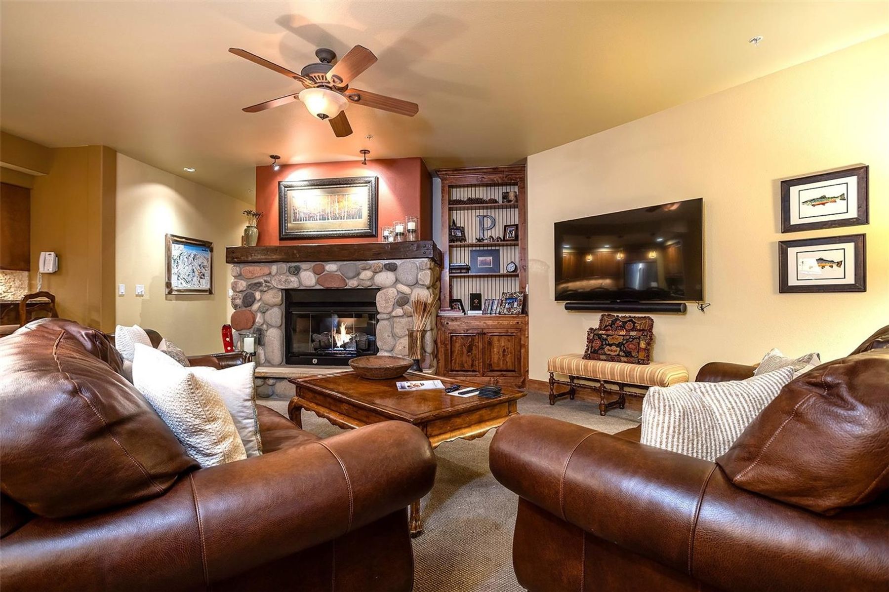 This cozy living room features a prominent stone-clad fireplace with a heavy wood mantel, serving as the focal point of the space. The room is furnished with plush brown leather sofas, a wooden coffee table, and a built-in wooden shelving unit, creating a warm and inviting atmosphere. A large wall-mounted television and framed artwork add to the comfortable, well-appointed aesthetic of this residential interior.