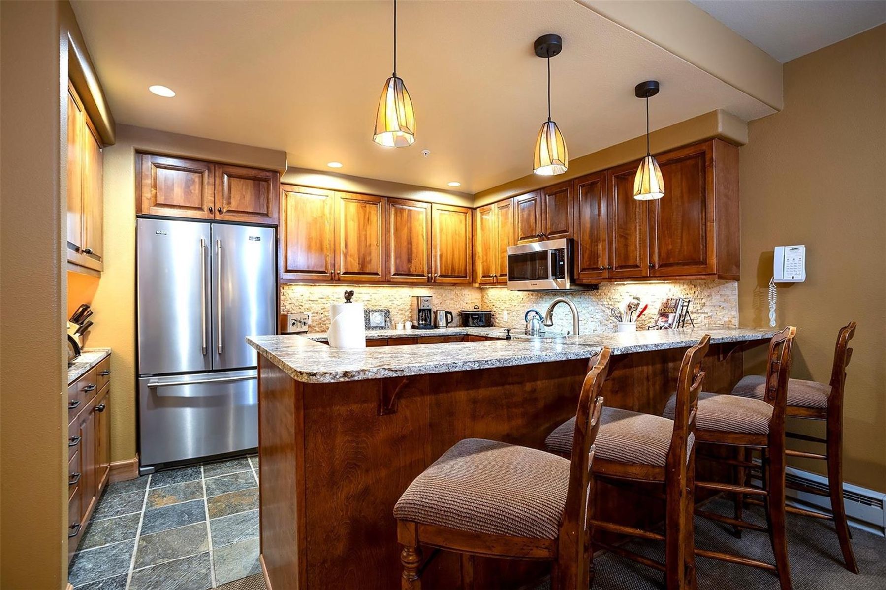 This warm and inviting kitchen features rich wood cabinetry, stainless steel appliances, and a granite-topped breakfast bar with three upholstered stools. The space is illuminated by three pendant lights hanging over the counter, complemented by under-cabinet lighting that highlights the textured stone backsplash. The floor is finished with large, multi-toned square tiles, creating a cohesive and functional culinary space.