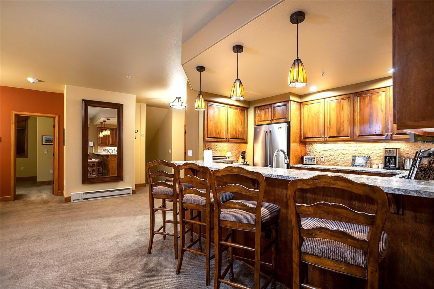 This kitchen features a breakfast bar with three wooden stools, set against a backdrop of warm-toned cabinetry and stone-look countertops. Three pendant lights hang above the counter, providing focused illumination over the workspace, while a large mirror on the adjacent wall reflects the room's layout. The space is carpeted and transitions into an open-concept living area, creating a cozy and inviting atmosphere.