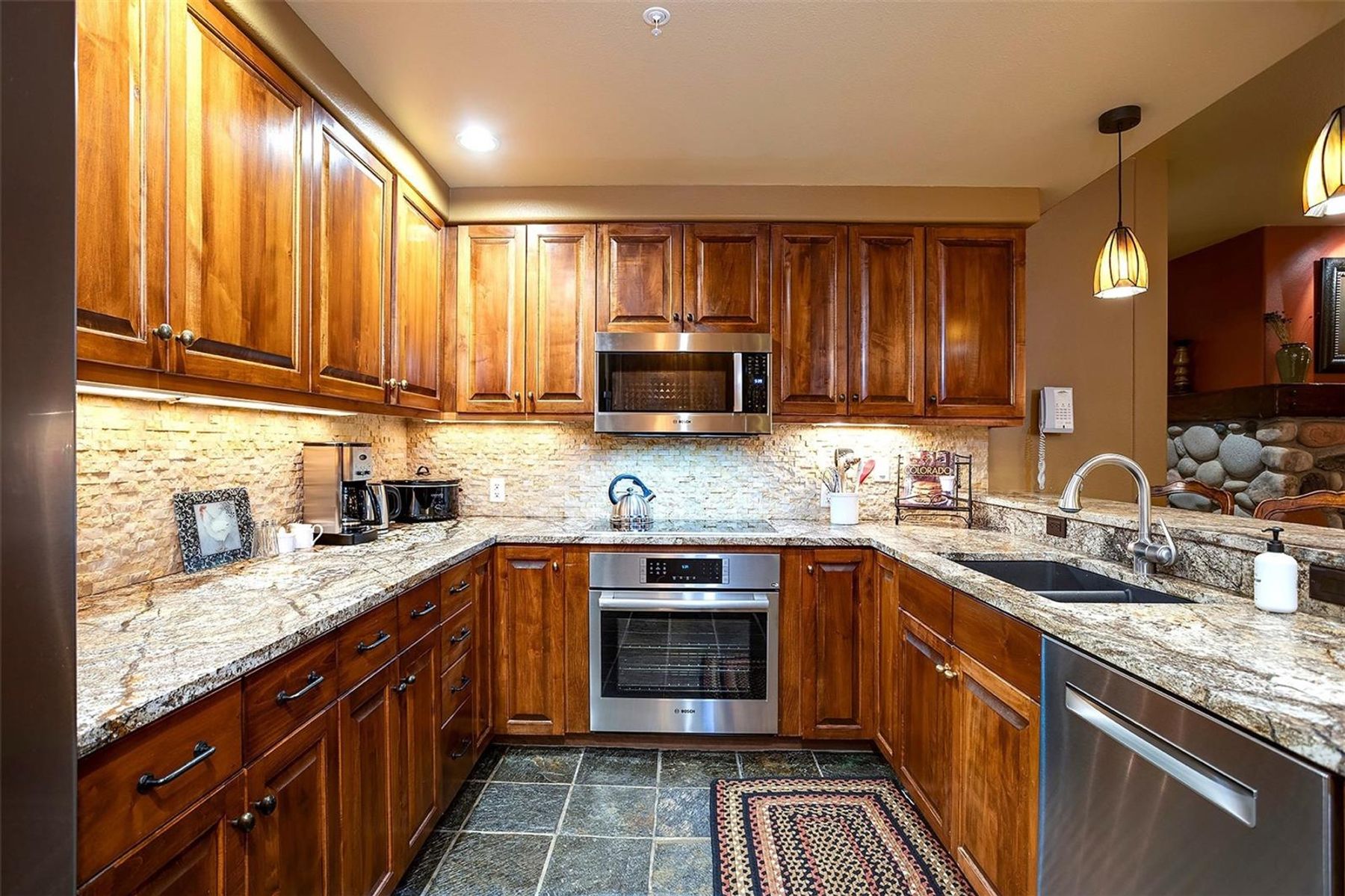 This U-shaped kitchen features warm-toned wood cabinetry paired with light-colored stone countertops and a textured stone backsplash. The space is equipped with stainless steel appliances, including a built-in oven and microwave, and is illuminated by under-cabinet lighting and pendant fixtures. A dark, patterned tile floor adds a rustic touch, while the open layout connects the cooking area to an adjacent living space with a stone-clad feature wall.