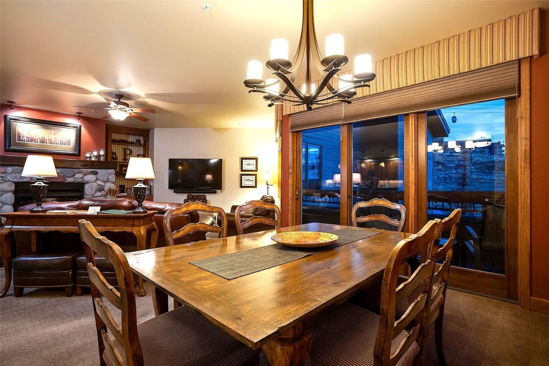 This dining area features a rustic wooden table centered beneath a multi-armed chandelier, creating a warm and inviting atmosphere. The space flows seamlessly into an adjacent living room, which is anchored by a stone-clad fireplace and comfortable leather seating. Large glass sliding doors provide a view of the exterior, while the open-concept layout emphasizes a cozy, mountain-lodge aesthetic.