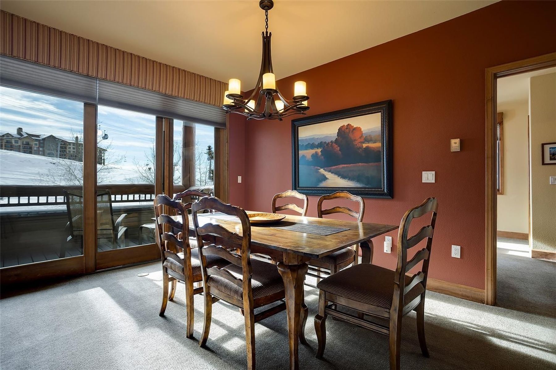 This dining room features a rustic wooden table surrounded by six matching chairs with curved backs and upholstered seats. A large, dark-framed landscape painting hangs on the deep red wall, while a distinctive antler-style chandelier provides overhead lighting. Large glass sliding doors offer a clear view of a snowy outdoor landscape, filling the room with natural light.