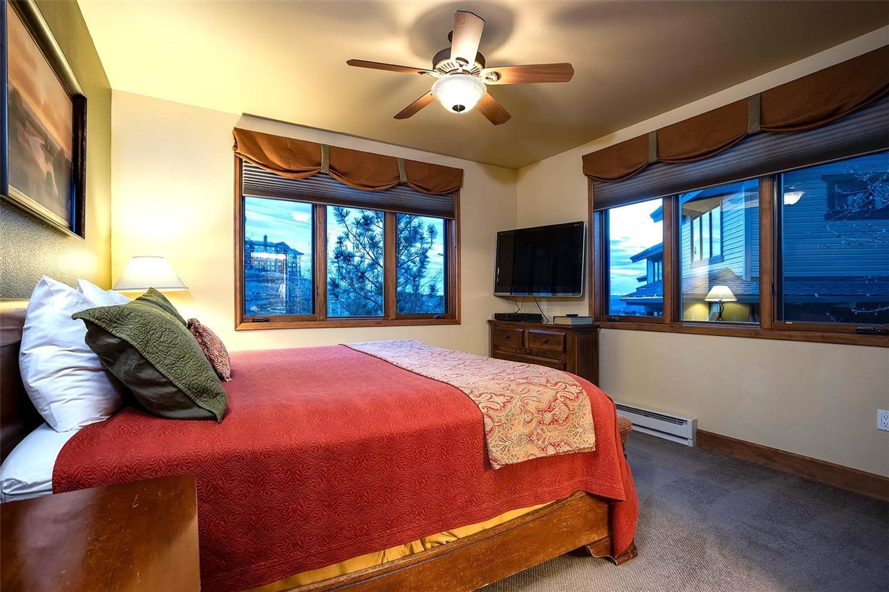 This cozy bedroom features a large bed with a vibrant red quilt and patterned accent runner, positioned centrally in the room. The space is framed by two sets of windows with brown valances that offer views of the surrounding landscape, while a ceiling fan provides overhead lighting. A wooden dresser sits between the windows, supporting a flat-screen television, creating a functional and comfortable retreat.
