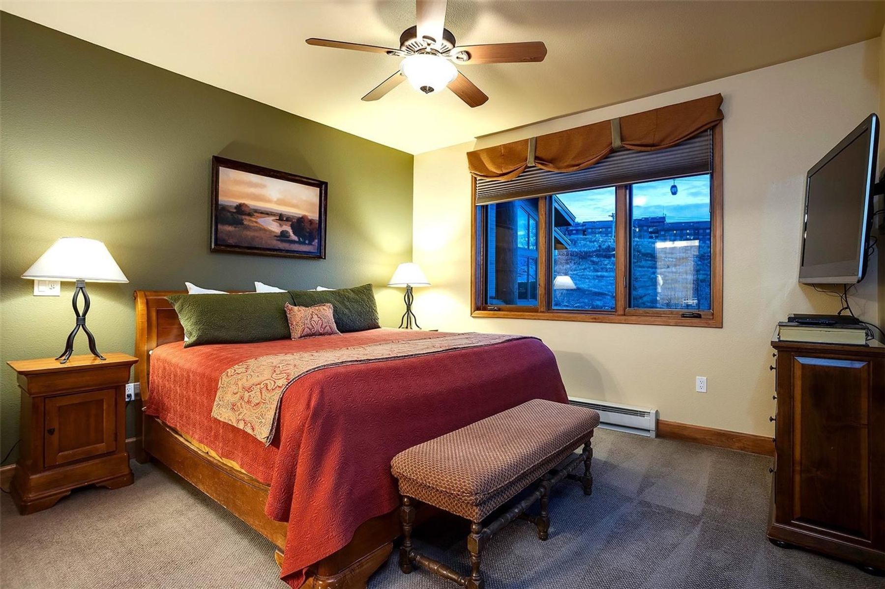 This cozy primary bedroom features a warm, rustic aesthetic with an olive-green accent wall and a large bed dressed in deep red bedding. A wooden nightstand with a lamp sits on either side of the bed, while a matching wooden dresser supports a television on the right. A large window with a fabric valance provides a view of the outdoors, and a cushioned bench is positioned at the foot of the bed.