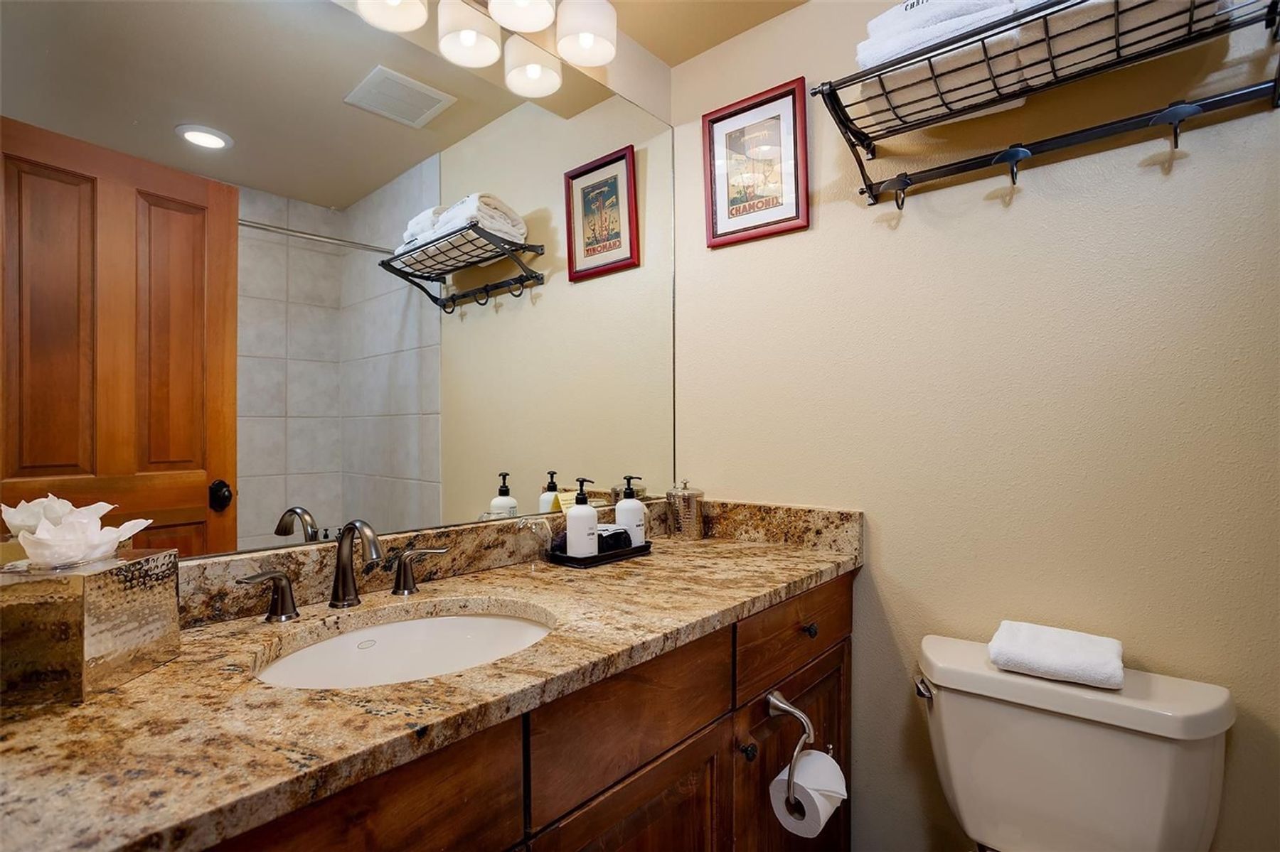 This bathroom features a warm, rustic aesthetic with a wood vanity topped by a speckled stone countertop and an undermount sink. A large mirror spans the wall above the vanity, reflecting the shower area and two framed vintage-style prints. The space is completed by a neutral-toned toilet, wall-mounted towel racks, and a multi-bulb light fixture, creating a cozy and functional atmosphere.