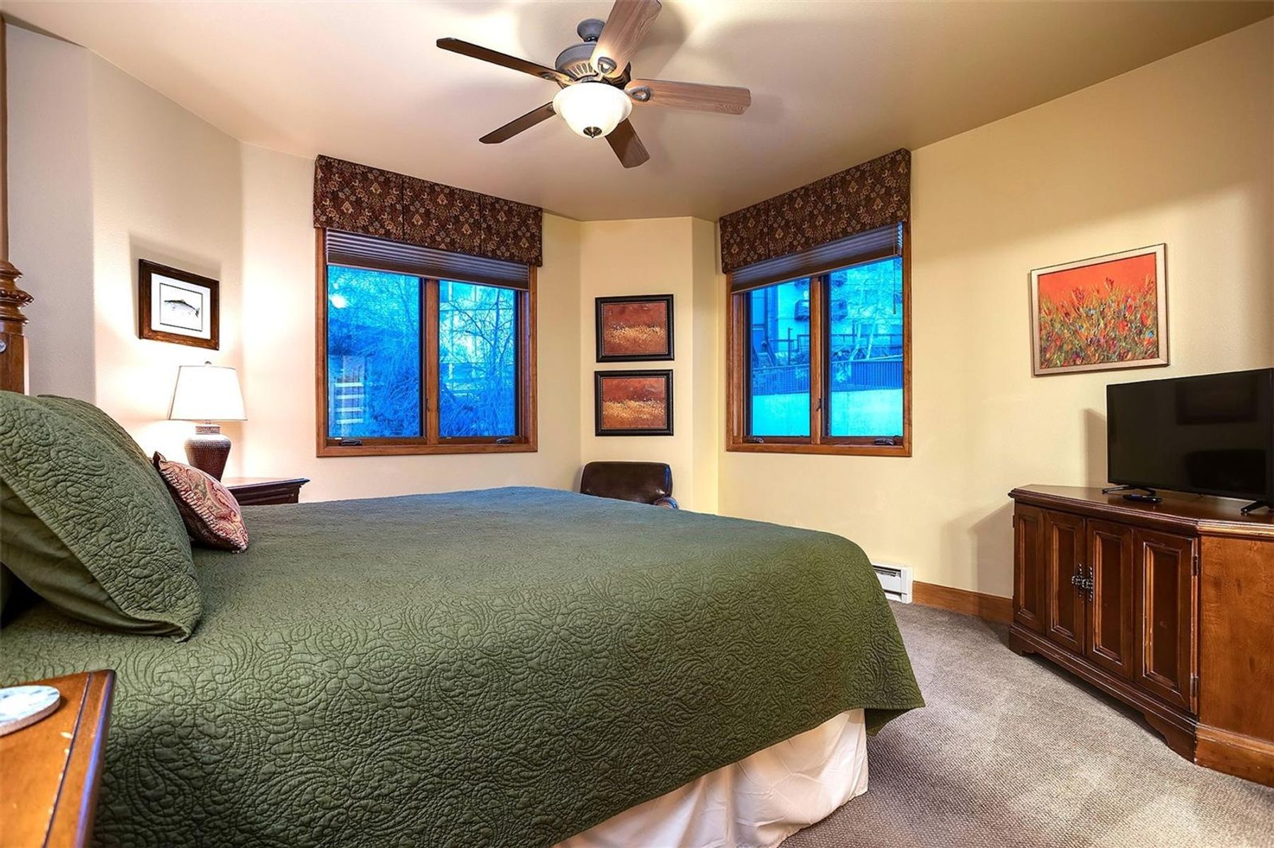 This cozy bedroom features a large bed with a textured green quilt, positioned centrally in the room. The space is illuminated by a ceiling fan with integrated lighting and a bedside lamp, while two windows with patterned valances provide natural light. A wooden dresser with a television sits to the right, and the room is decorated with framed artwork on the walls, creating a warm and inviting atmosphere.