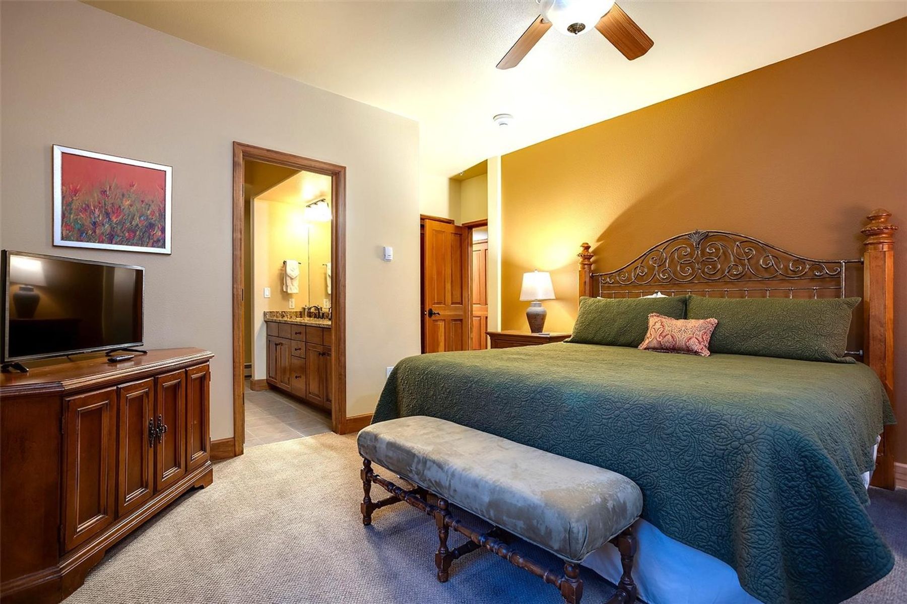 This spacious primary bedroom features a large bed with a decorative metal headboard, dressed in a dark green quilted coverlet and a patterned accent pillow. A wooden dresser with a television sits to the left, while a cushioned bench rests at the foot of the bed, and an open doorway leads into an adjoining bathroom. The room is accented by a warm, golden-toned feature wall and soft overhead lighting from a ceiling fan, creating a cozy and inviting atmosphere.