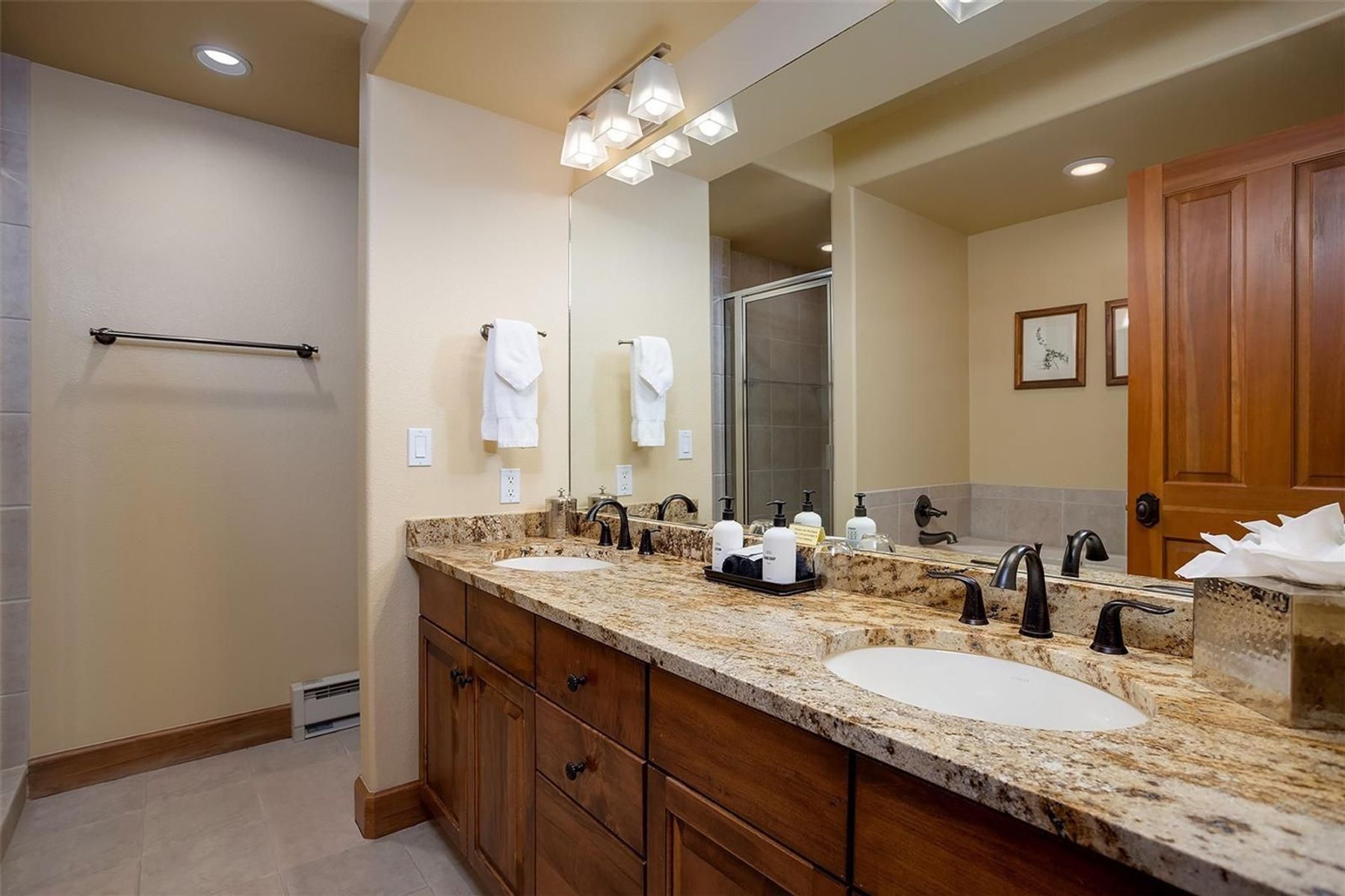 This primary bathroom features a dual-sink vanity with a warm-toned wood finish and a speckled stone countertop. A large, full-width mirror reflects the space, including a glass-enclosed shower and a wooden door leading to the rest of the home. The room is illuminated by a multi-bulb vanity light fixture and recessed ceiling lighting, creating a bright and functional atmosphere.