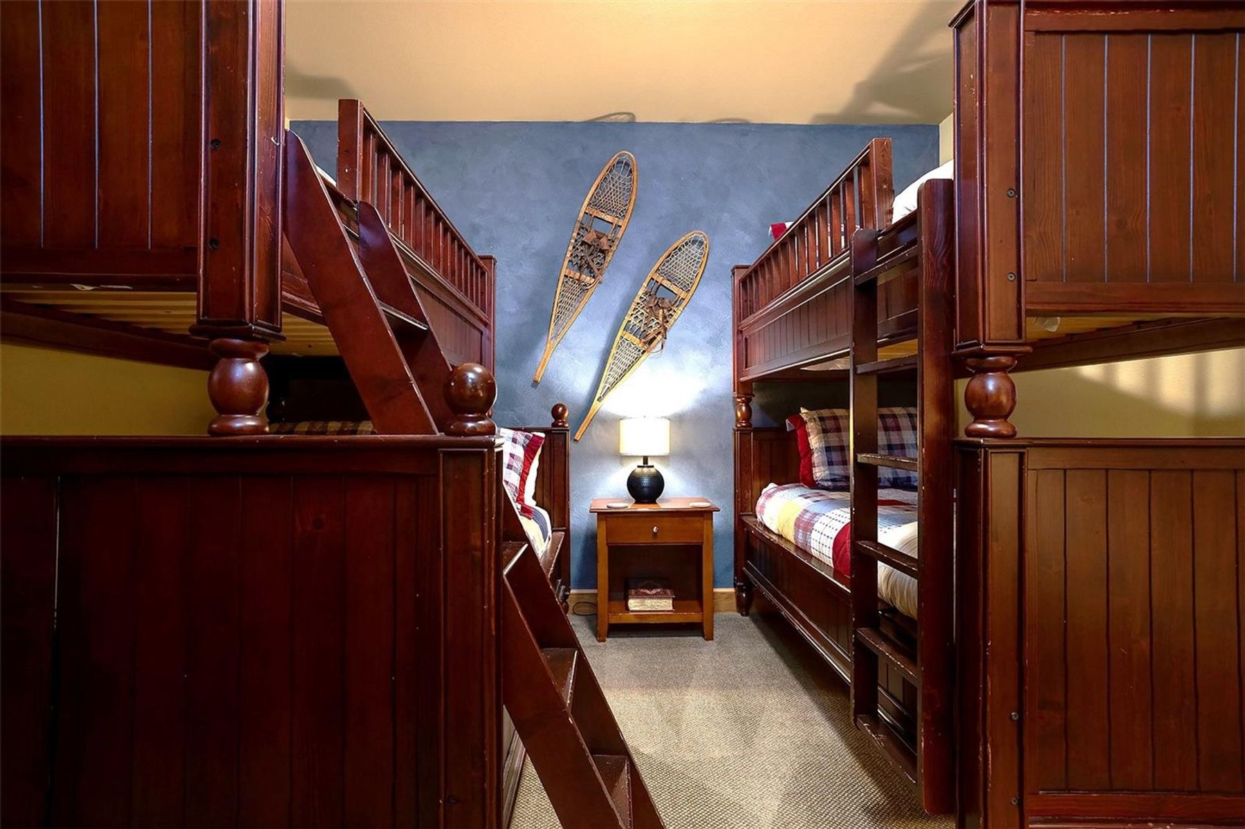 This cozy bedroom features two sets of dark wood bunk beds positioned on either side of a central nightstand, creating a symmetrical and efficient sleeping space. The walls are painted a deep blue, accented by decorative wooden snowshoes mounted above the lamp, which adds a rustic, lodge-inspired aesthetic. The room is carpeted and illuminated by a warm, soft glow from the bedside lamp, emphasizing a comfortable and inviting atmosphere.
