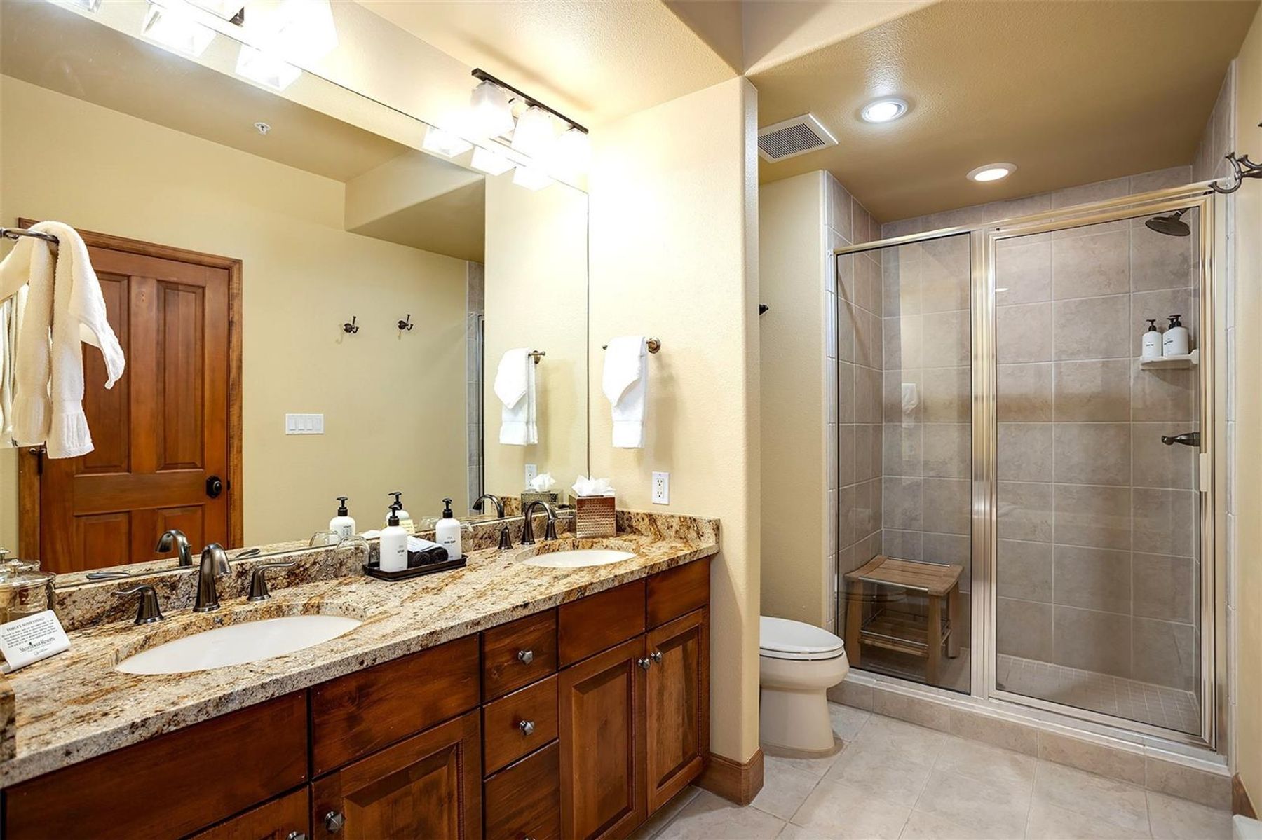 This primary bathroom features a dual-sink vanity with a stone countertop and dark wood cabinetry, complemented by a large mirror and bright vanity lighting. To the right, a glass-enclosed walk-in shower includes a built-in wooden bench and neutral-toned wall tiles. The space is finished with light-colored floor tiles and a neutral wall color, creating a clean and functional atmosphere.