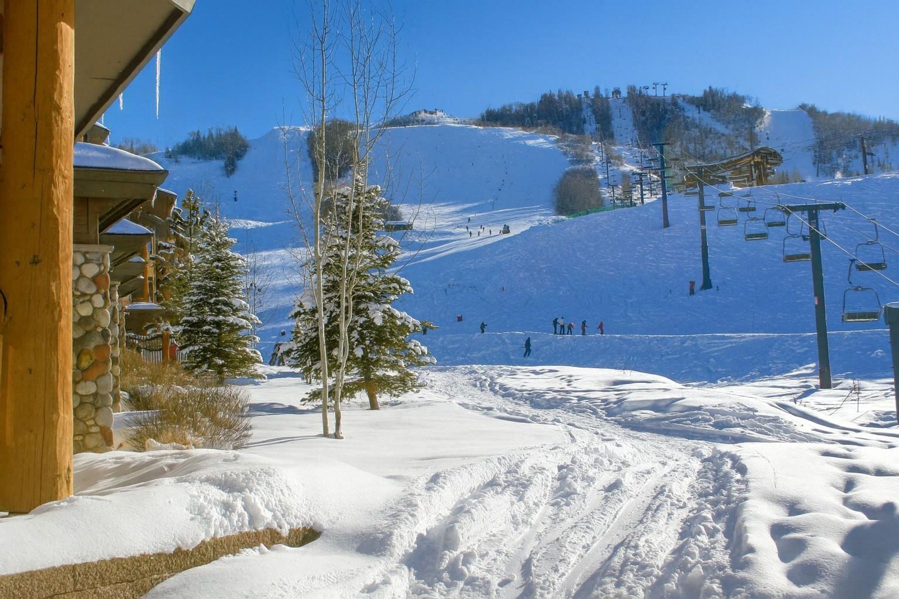 This view from a covered patio or balcony captures a prime ski-in/ski-out location at a mountain resort. The foreground features a rustic timber-framed structure with stone accents, while the background showcases a snow-covered ski slope with a chairlift and skiers in action. The bright, clear blue sky and pristine snow create an inviting, high-altitude atmosphere perfect for winter sports enthusiasts.