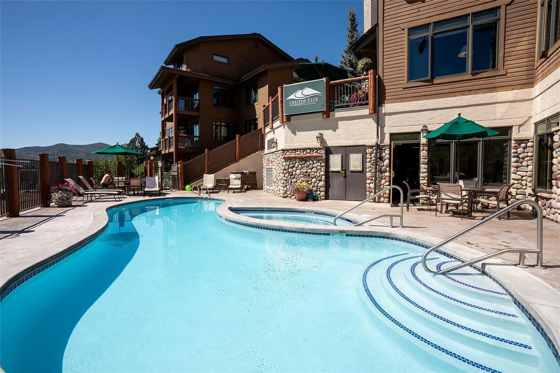 This outdoor amenity area features a curved swimming pool with integrated steps and a separate adjacent spa. The pool deck is surrounded by lounge chairs, green umbrellas, and stone-accented building exteriors, creating a resort-like atmosphere. The scene is captured from a ground-level perspective, highlighting the inviting water and the mountain backdrop under a clear blue sky.