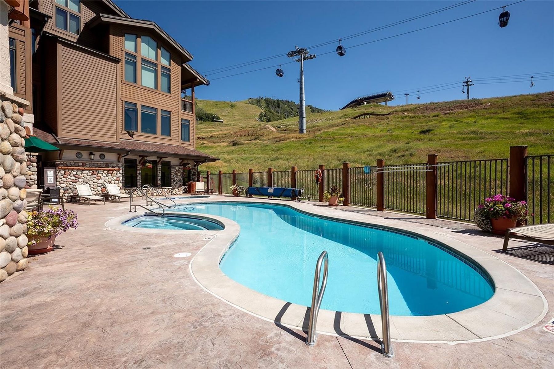 This outdoor space features a kidney-shaped swimming pool and an integrated hot tub set on a stamped concrete patio. The area is bordered by a metal fence that overlooks a grassy hillside with a ski lift system visible in the background. The adjacent building is constructed with brown siding and stone accents, creating a rustic mountain resort aesthetic.