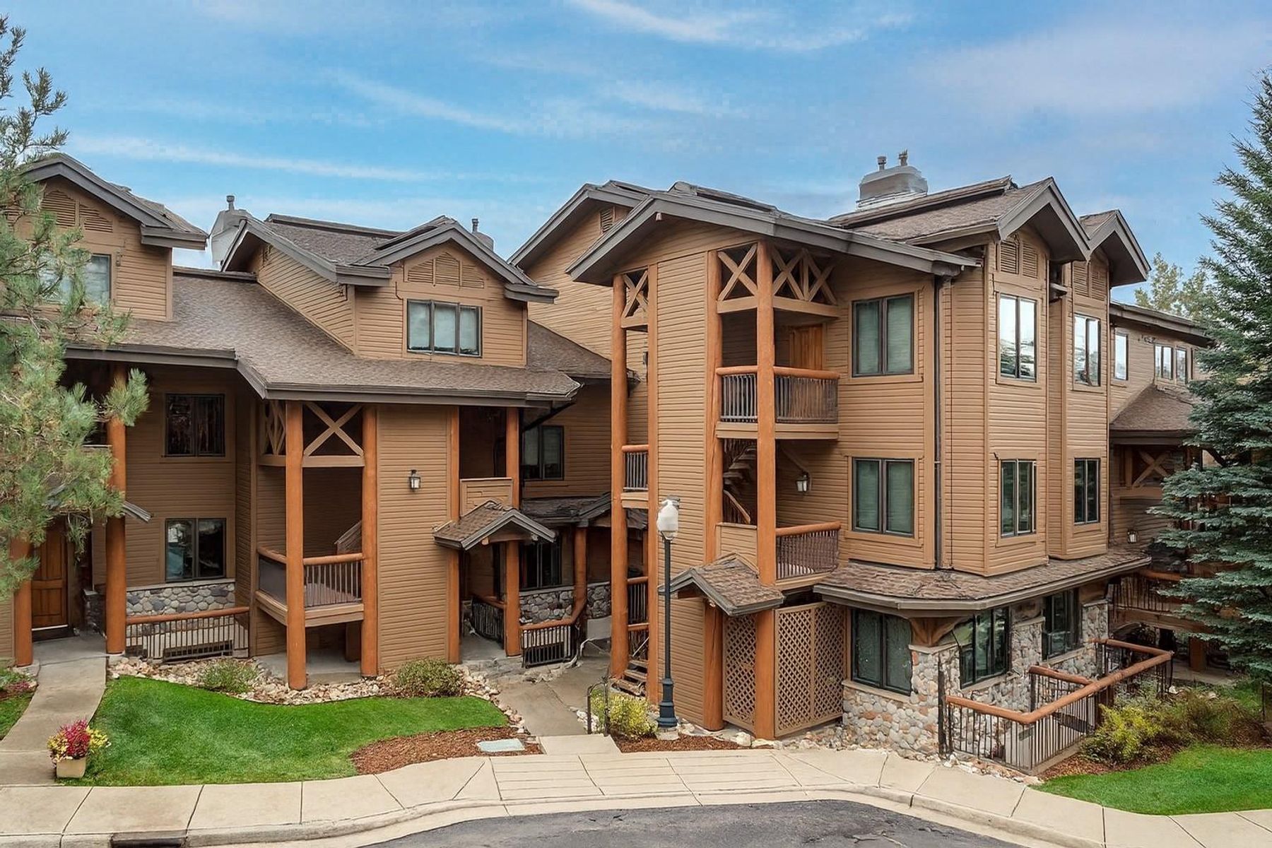 This multi-story residential building features a rustic mountain-inspired design with brown horizontal siding, stone accents, and dark shingled roofs. The structure includes multiple balconies with wooden railings, decorative timber framing, and a paved walkway leading to the main entrance. The scene is set against a clear blue sky with a manicured lawn and mature evergreen trees in the foreground.