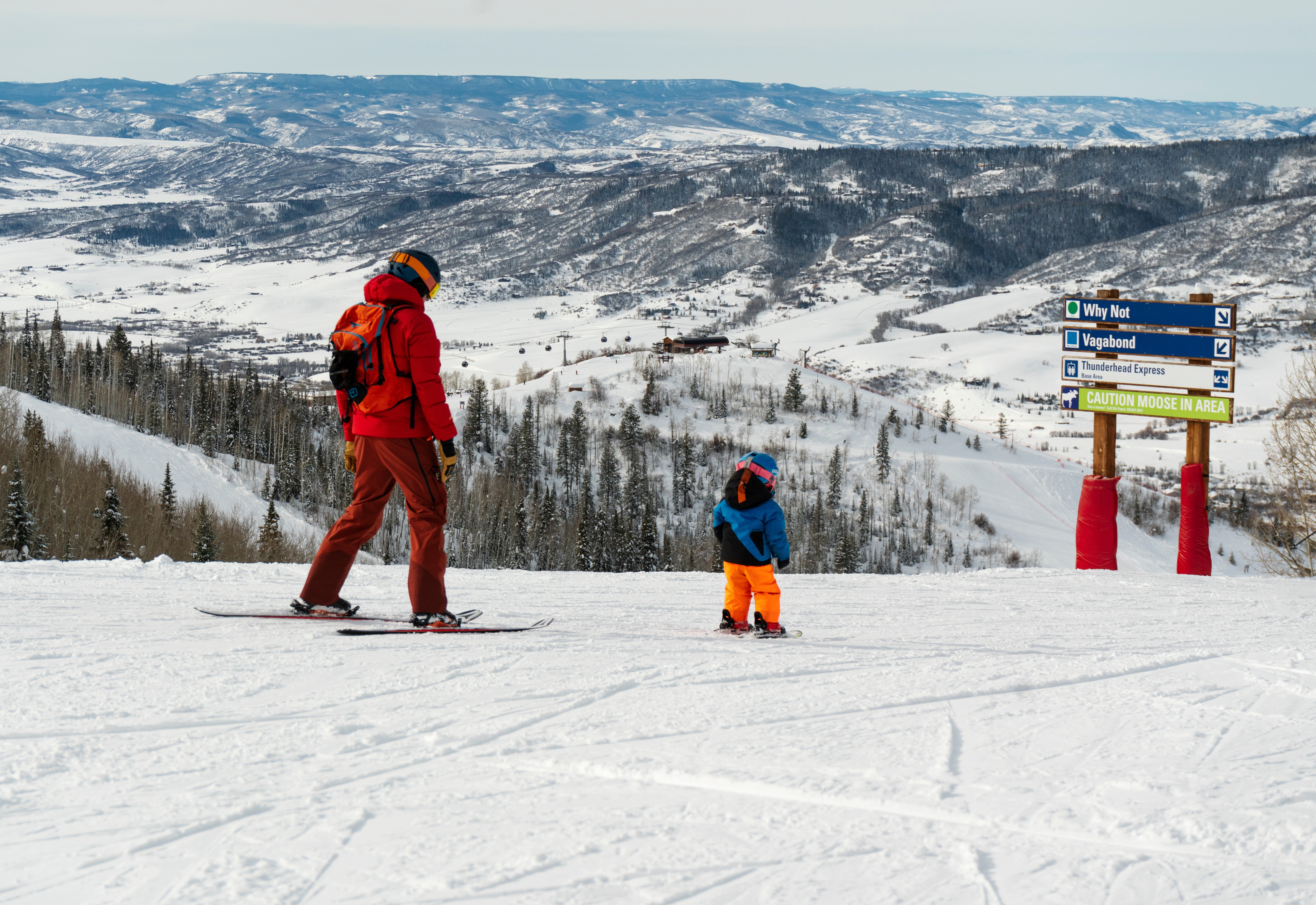 A wide-angle, eye-level shot captures a skier and a young child on skis standing on a snowy mountain ridge, overlooking a vast, expansive valley. The scene highlights the recreational lifestyle of a mountain resort, featuring a prominent trail sign indicating nearby runs and a caution notice for wildlife. The backdrop showcases a panoramic view of snow-covered peaks and forested slopes, emphasizing the scenic beauty and outdoor amenities of the location.