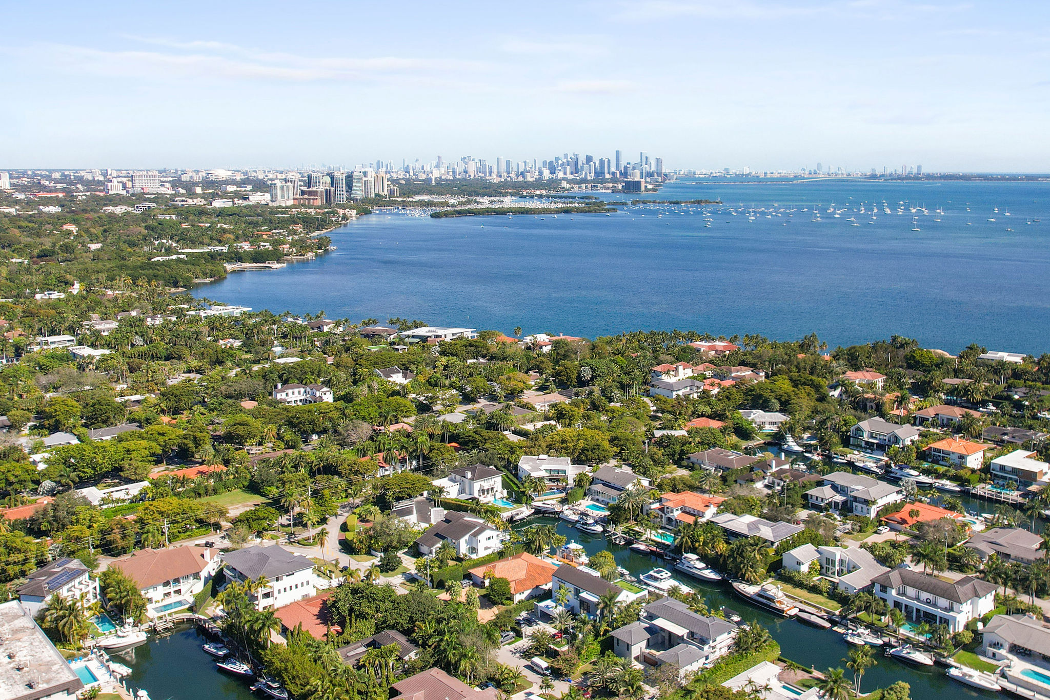 This aerial view showcases a luxurious waterfront community with upscale homes, private boat docks, and lush landscaping. Many homes feature pools and access to canals, appealing to boating enthusiasts. The distant cityscape adds an urban backdrop to this serene residential area, suggesting proximity to city amenities.