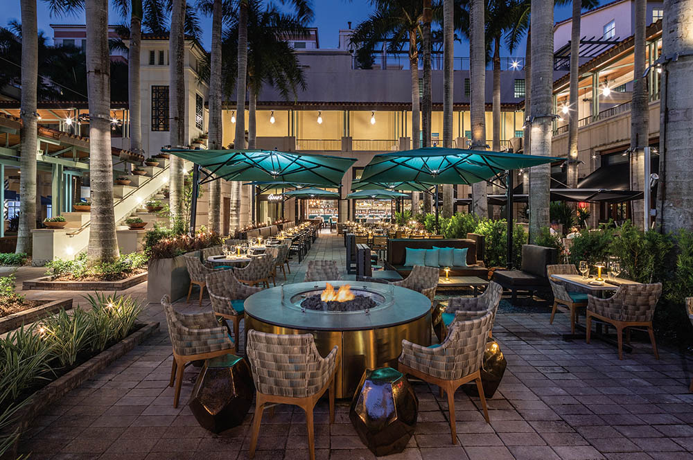 This is an inviting outdoor patio space featuring a fire pit surrounded by comfortable chairs. Several towering palm trees provide a tropical ambiance, while overhead teal umbrellas offer shade. The setup suggests a relaxing and upscale dining or lounge area, perfect for evening entertainment.