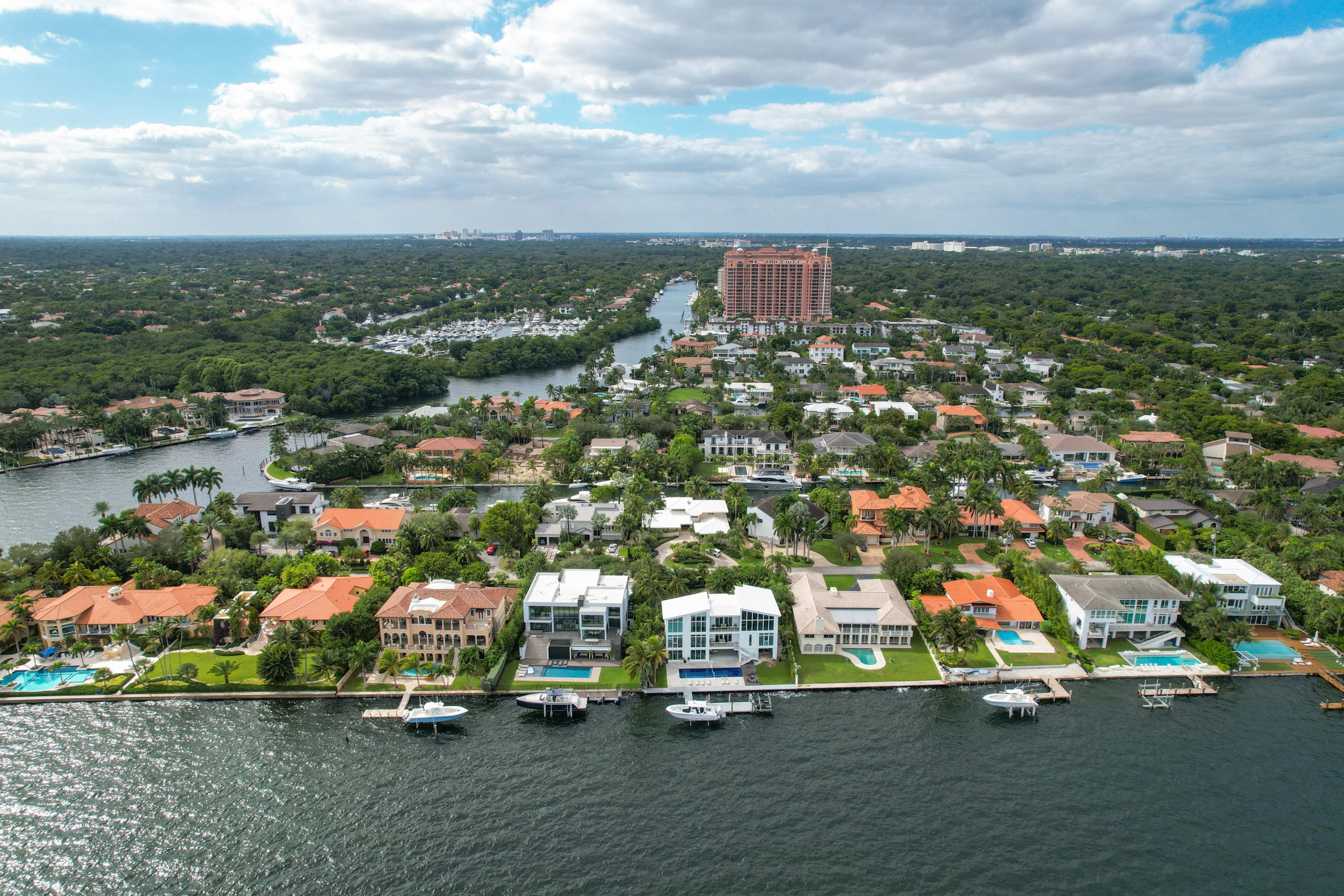 This aerial real estate image showcases waterfront properties with stunning architecture, featuring modern and Mediterranean-style homes. Swimming pools, private docks with boats, and lush landscaping are visible, adding to the appeal of these luxury homes. The scene is set against a backdrop of canals and dense greenery typical of upscale residential areas.