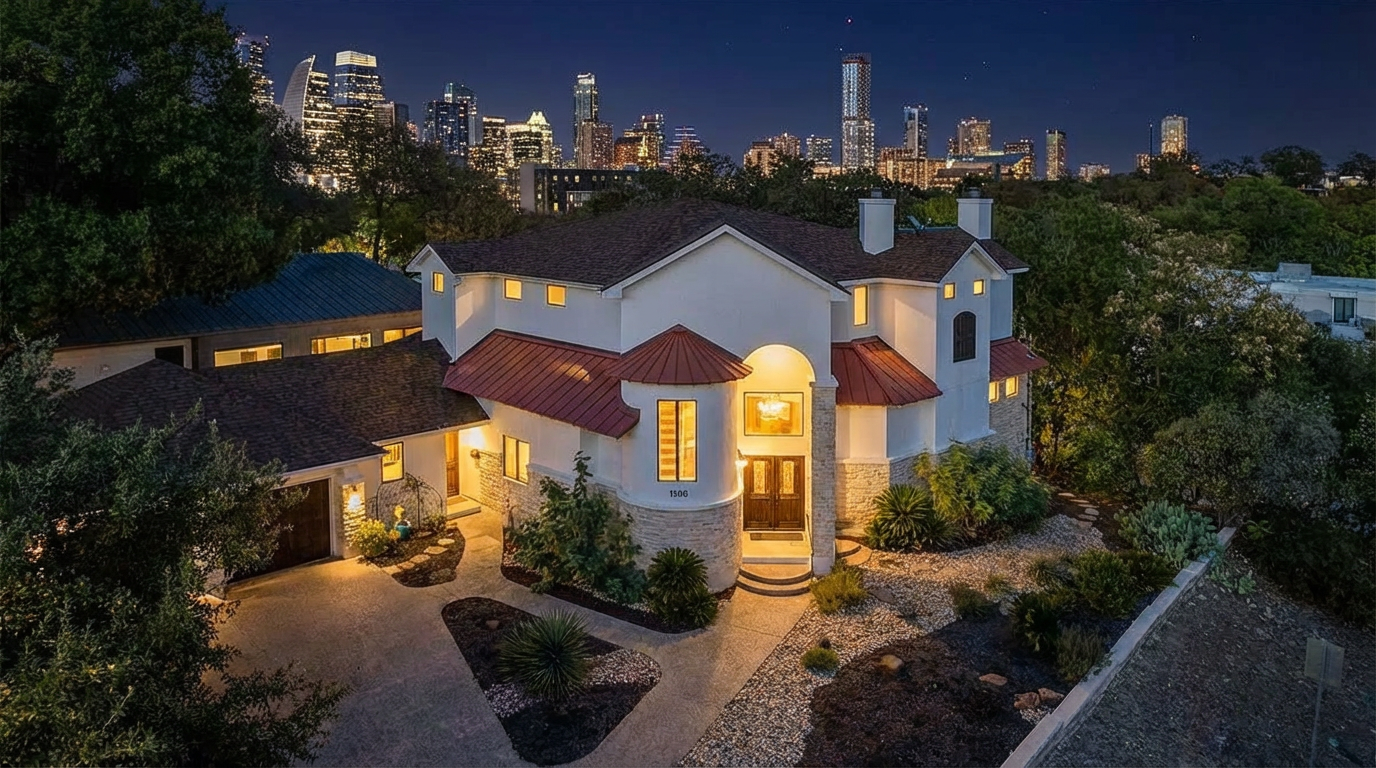 This aerial view showcases a large, luxurious home with a unique architectural style. The property features a well-manicured front yard and is situated in a location with a stunning skyline view in the background. The well-lit exterior highlights the home's features, making it an attractive real estate listing.