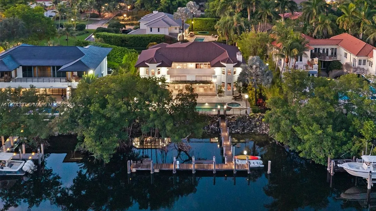 This aerial shot showcases a luxurious waterfront property with a large house, private dock, and boat access. The manicured landscaping and surrounding palm trees add to the estate's appeal. The residence features a clay tile roof and multiple balconies, creating a sophisticated and grand impression.