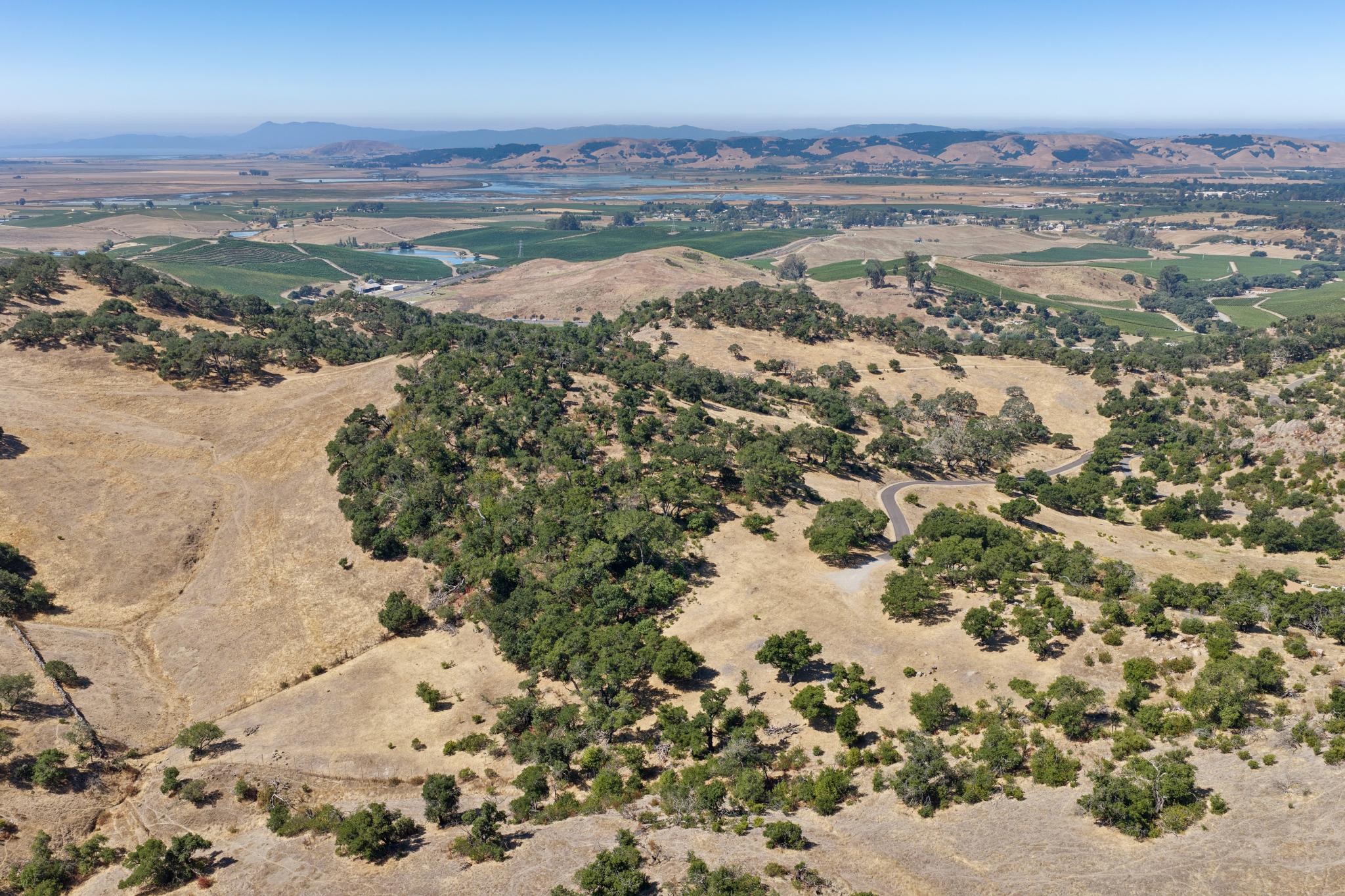 This stunning aerial photograph captures a expansive, rural landscape characterized by rolling hills, scattered oak trees, and dry, golden grasses. A winding paved driveway snakes through the terrain, suggesting private access to a secluded property nestled within this natural topography. The background features a picturesque valley with bodies of water and distant mountains, showcasing an idyllic, peaceful setting ideal for those seeking privacy and nature.