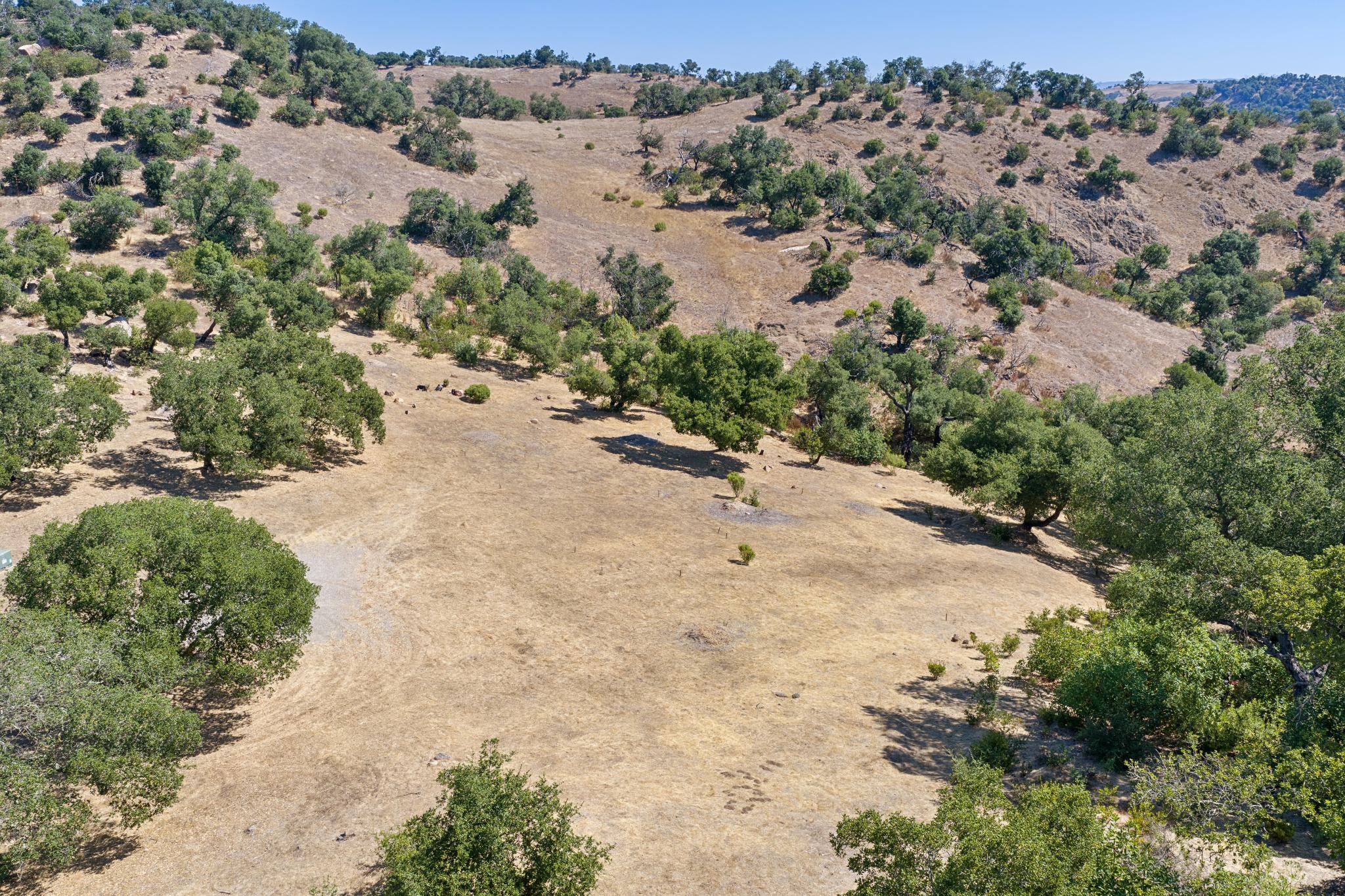 This high-angle aerial view showcases a expansive, sun-drenched rural landscape characterized by rolling hills covered in dry, golden grasses and scattered oak trees. The property features an open meadow area that offers significant potential for development or recreation. It captures the natural, rugged beauty of the terrain and provides a sense of the vast, private acreage available.