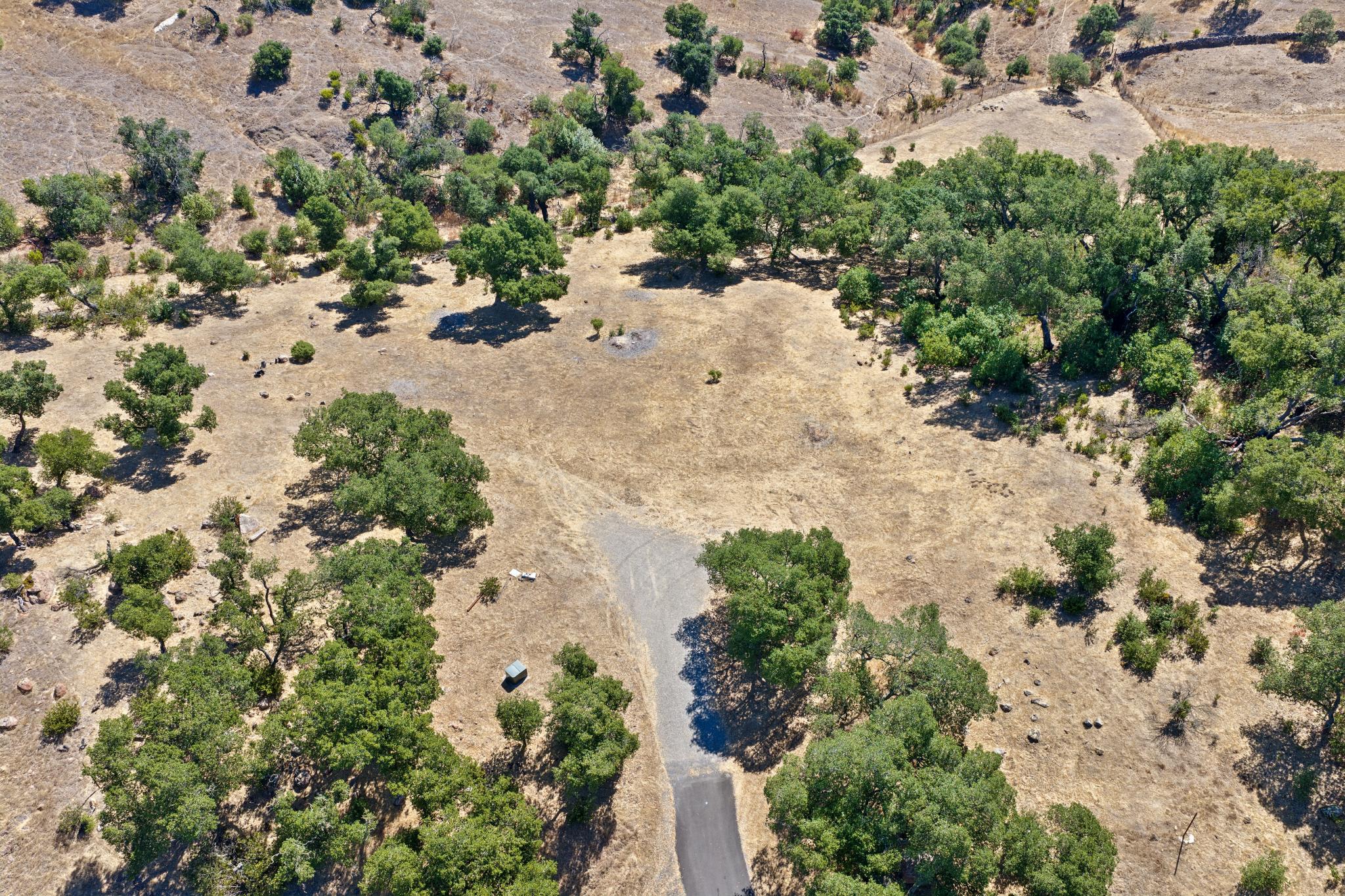 This aerial view showcases a spacious, undeveloped plot of land characterized by rolling, dry terrain dotted with numerous oak trees. A paved driveway leads into the property, providing clear access to a potential building site. The landscape offers a sense of privacy and natural beauty, ideal for those seeking a tranquil, rural retreat.