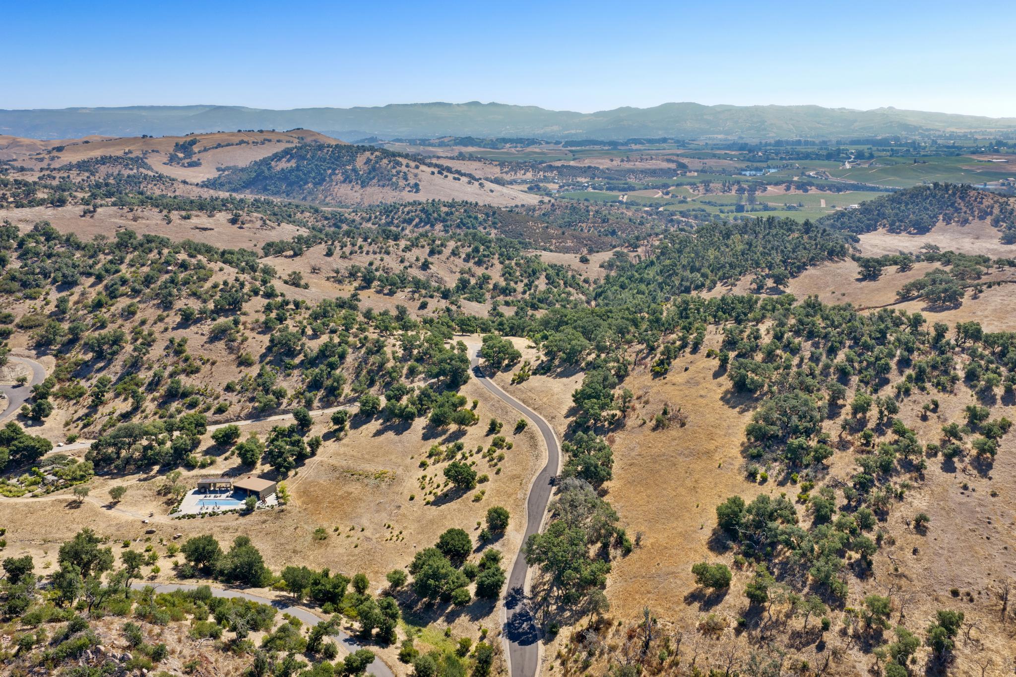 This aerial view captures a sprawling, rural estate set against a backdrop of rolling hills and distant mountains. A long, winding driveway leads to a modern residence equipped with a swimming pool, showcasing a private and serene landscape characterized by scattered oak trees and open meadows. The setting highlights the expansive nature of the property and its integration into the natural California terrain.