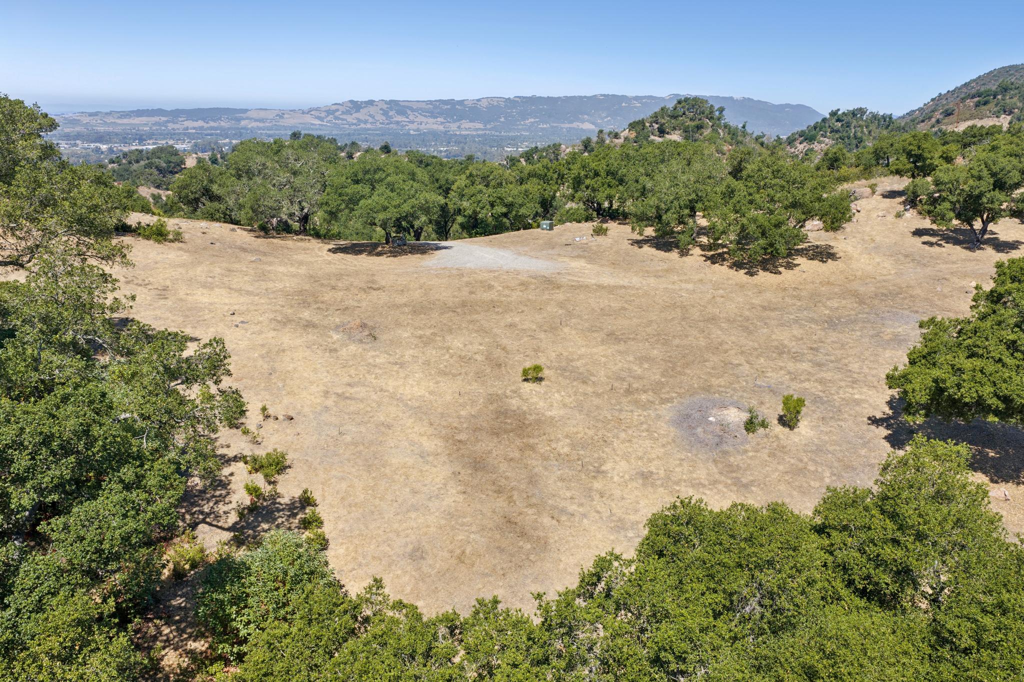 This high-angle aerial view showcases a expansive, sun-drenched plot of undeveloped land characterized by dry, golden grasses and mature oak trees. The property features a clear, elevated vantage point overlooking distant mountains and valley scenery, making it a prime candidate for custom residential construction. The scene highlights the natural terrain and peaceful, secluded atmosphere of the rural landscape.