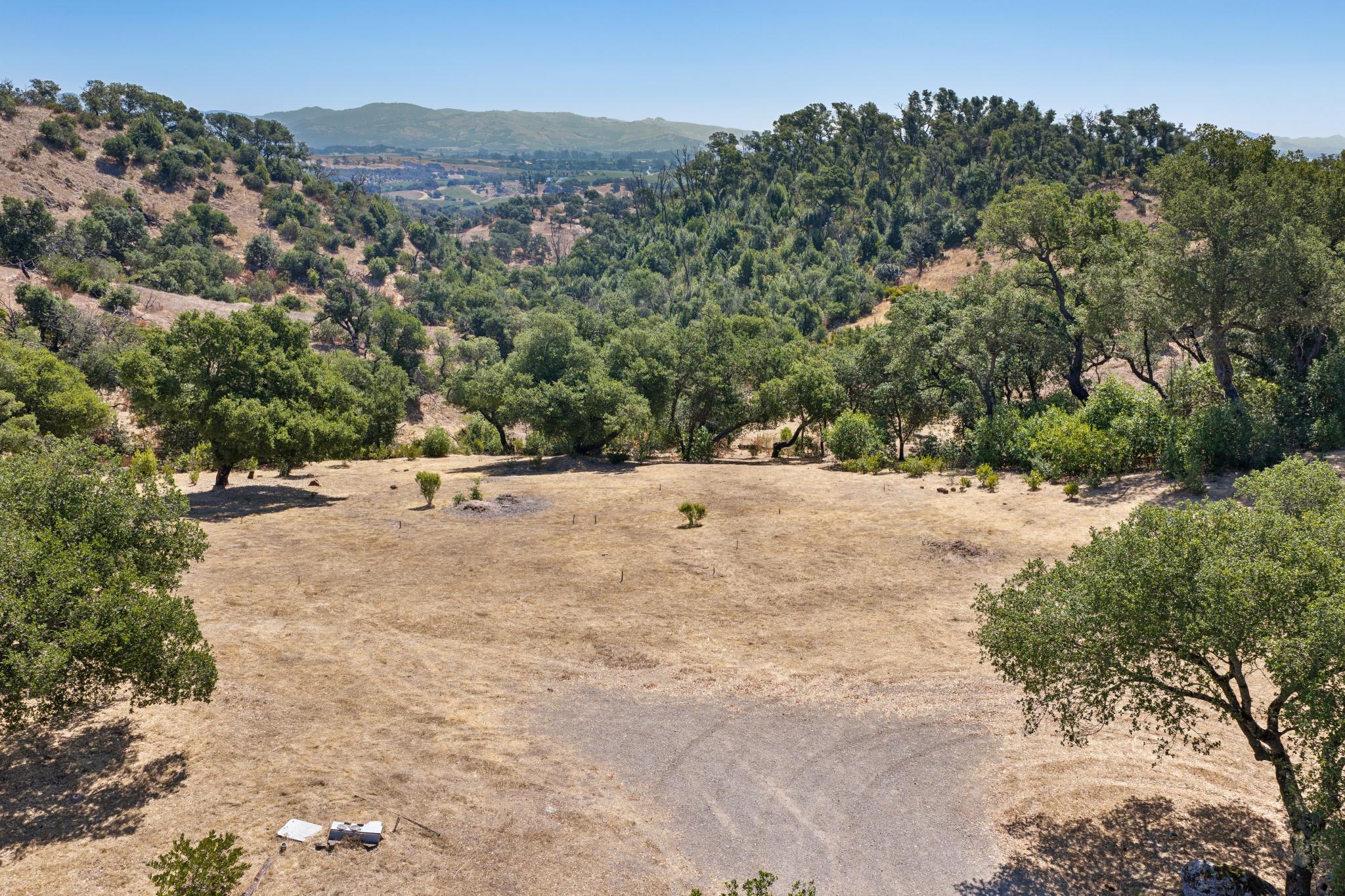 This high-angle, aerial perspective showcases a sprawling, rural residential lot characterized by dry, golden-hued pasture land. The property features mature oak trees scattered across the terrain and transitions into a lush, forested hillside backdrop that offers scenic views of the distant valley.