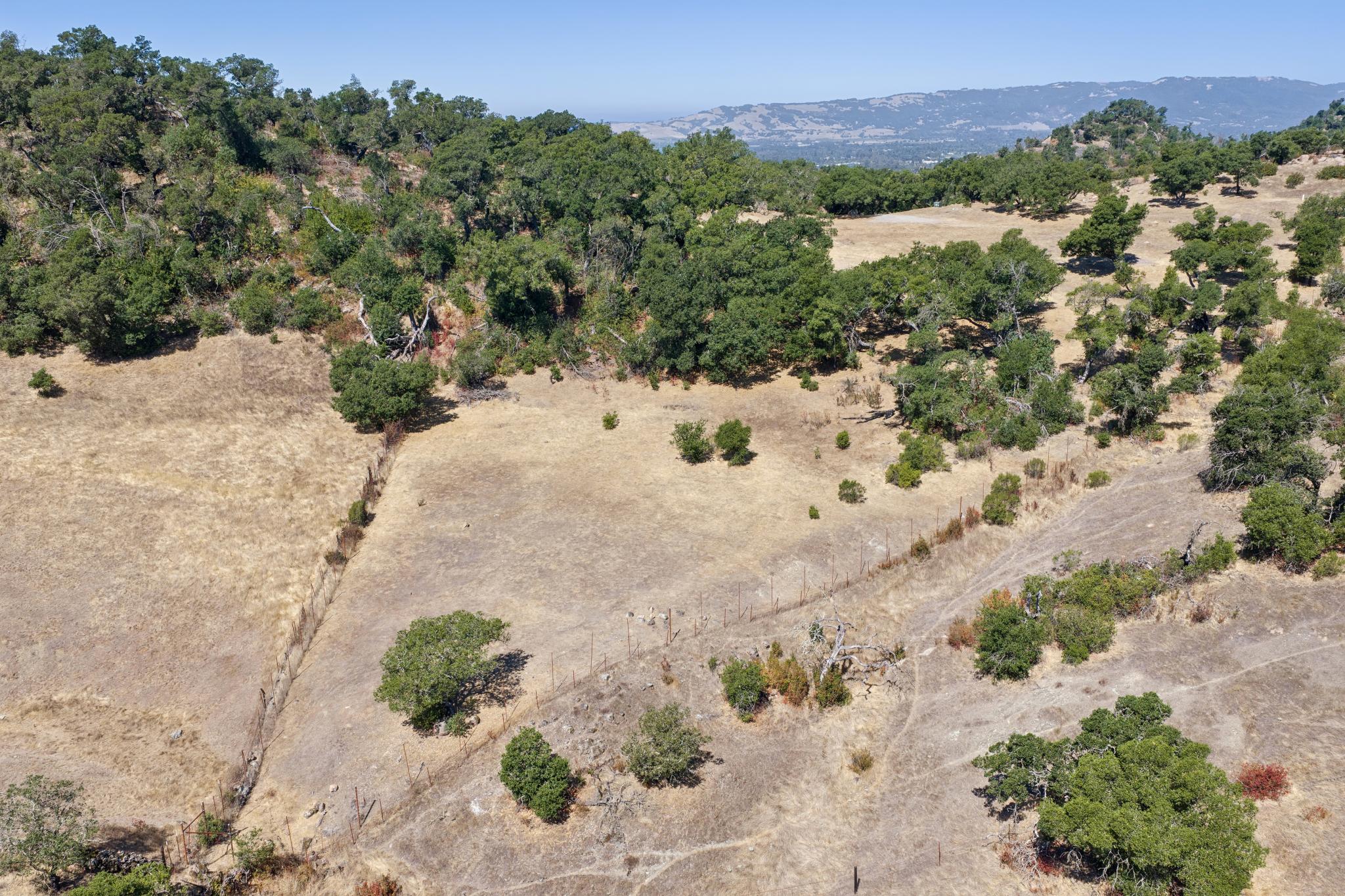 This aerial view showcases a sprawling, hilly landscape characterized by dry, golden grassy slopes and numerous oak trees. The property features natural terrain ideal for outdoor activities, with a rustic wire fence demarcating portions of the land, providing a sense of space and seclusion.