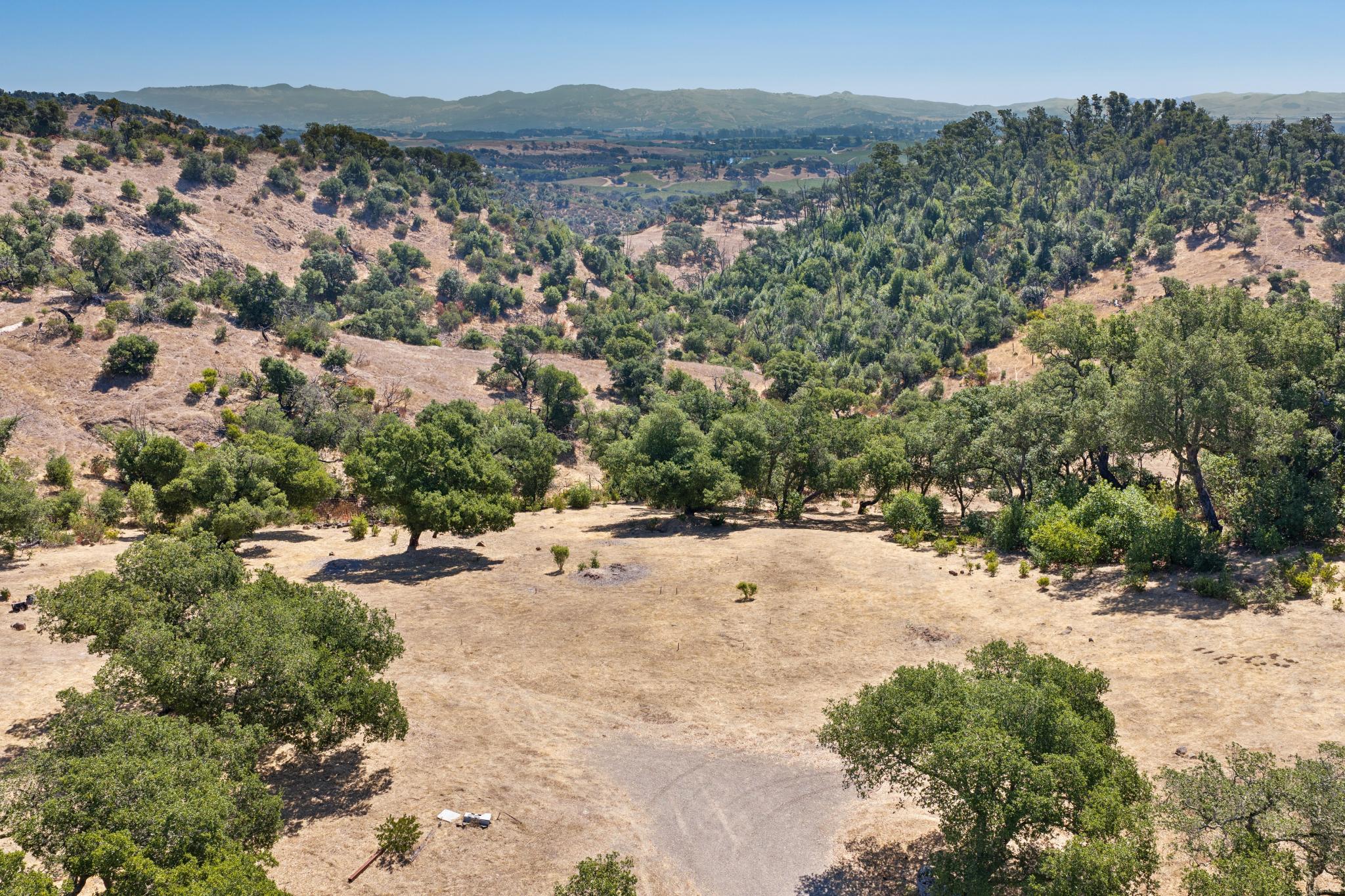 This wide-angle aerial perspective captures a sprawling rural landscape characterized by rolling, golden-hued hills and scattered mature oak trees. The property features a natural, rustic topography leading into a wooded valley, with distant mountain ranges providing a scenic backdrop. It offers an expansive sense of privacy and serene natural beauty, ideal for buyers seeking a secluded retreat.