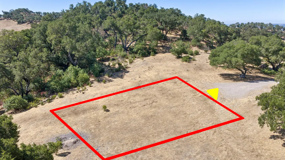 This aerial view showcases a cleared, rectangular lot outlined in red, set within a natural, wooded landscape. The dry, grassy terrain suggests a rural or semi-rural setting with mature oak trees bordering the property. The presence of a small gravel access point adds to the parcel's potential for future development.