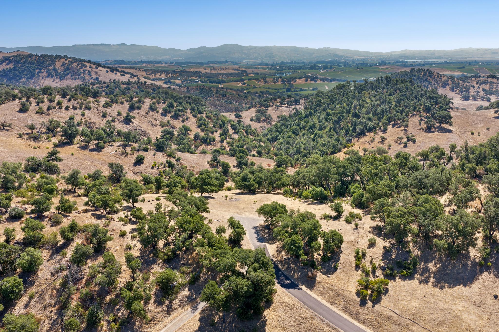 This high-altitude aerial view captures a sprawling, hilly landscape characterized by dry, golden grasses and clusters of mature oak trees. A paved driveway winds through the terrain, leading toward a secluded area that suggests potential for a residential build site. The scene offers expansive, scenic vistas of the surrounding valley and distant mountains, highlighting a peaceful, private, and rural setting.