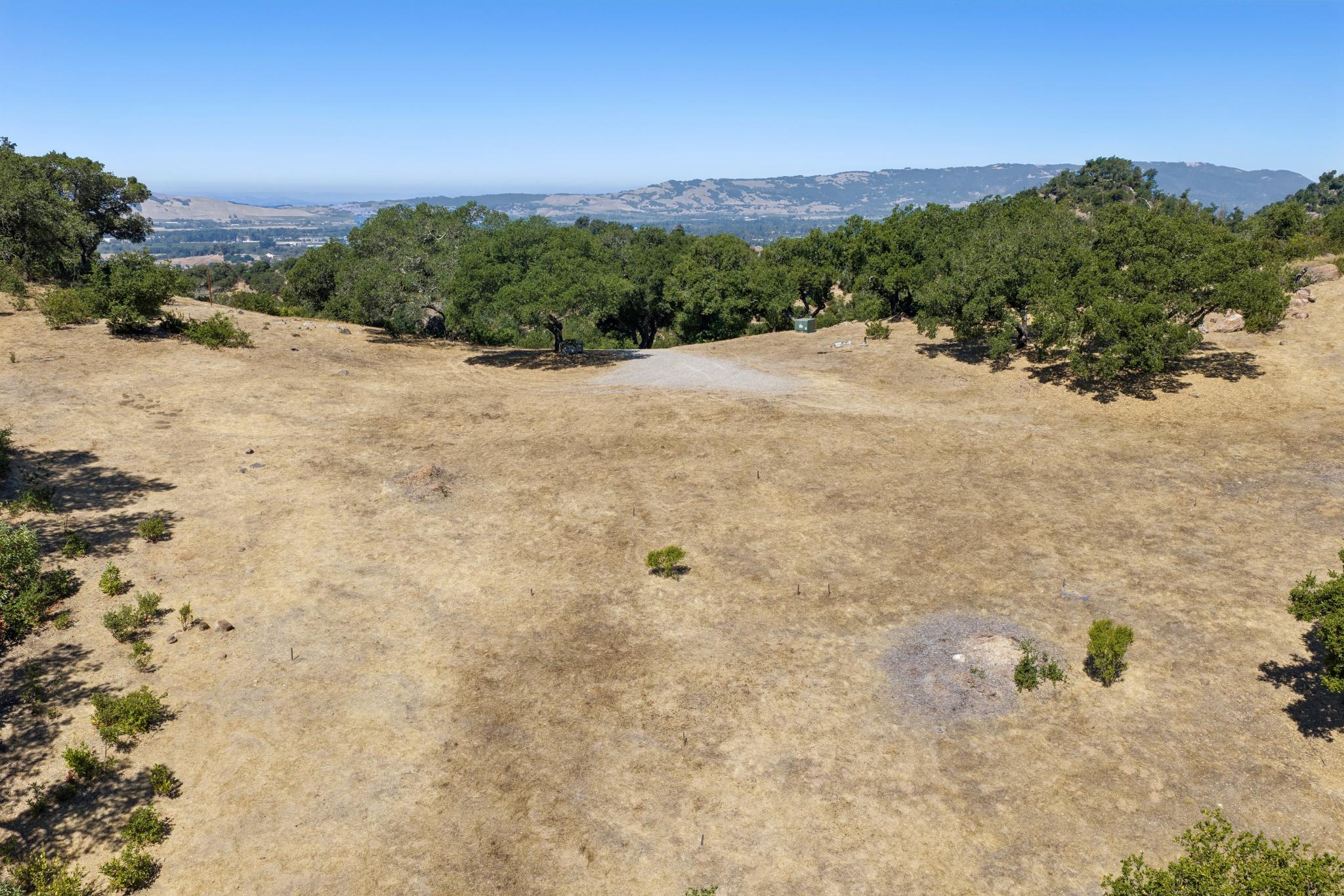 This elevated view captures an expansive, sun-drenched plot of land characterized by dry, golden grass and scattered oak trees. The property features a natural landscape with a gravel access area, suggesting a prime location for future development or a private retreat. The distant hills and clear blue sky provide a scenic backdrop, highlighting the potential for panoramic views.