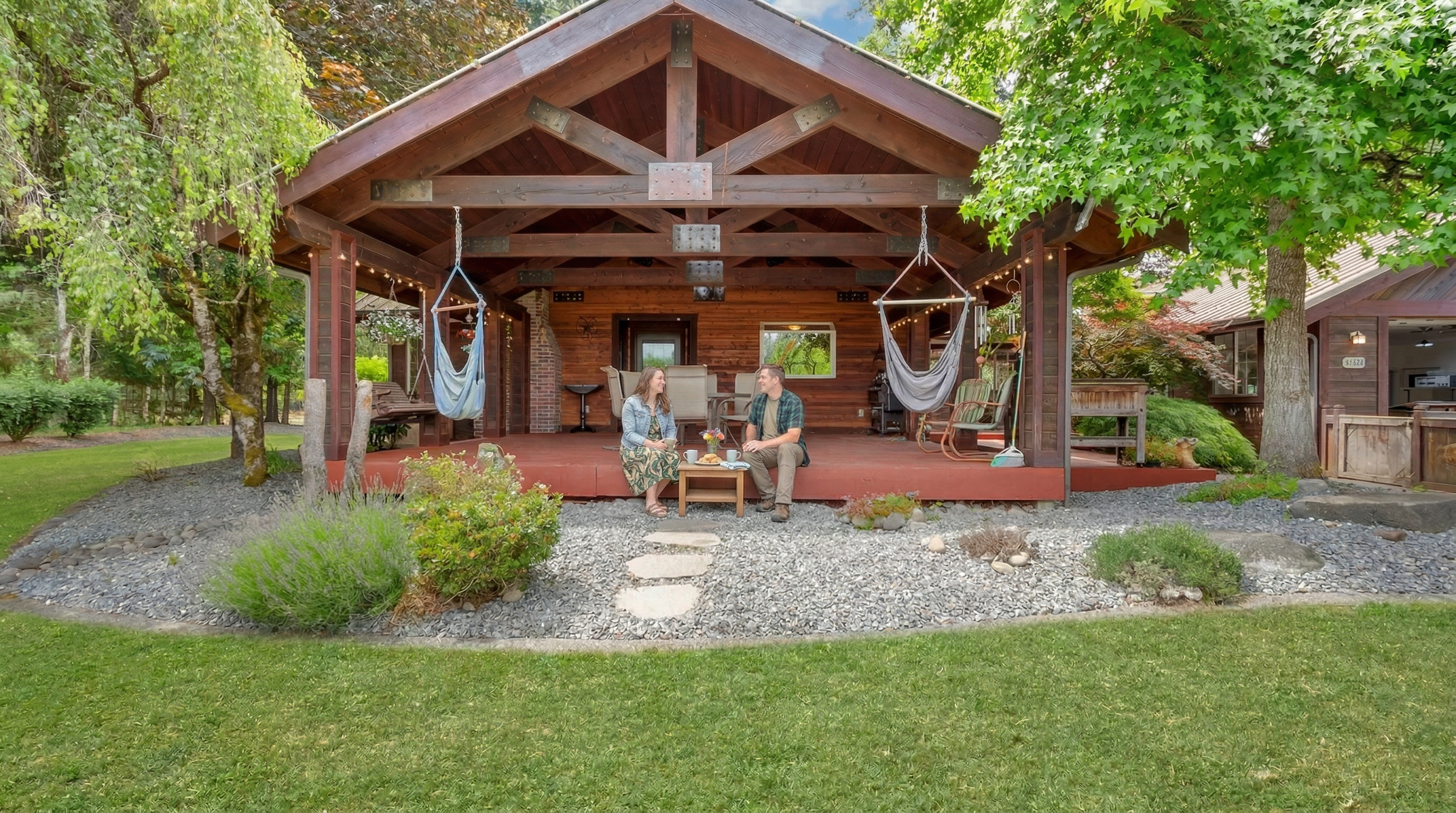 The image showcases an inviting outdoor patio or deck area. The structure features exposed wooden beams and supports, creating a rustic and cozy atmosphere. Comfortable seating arrangements including chairs and hammocks are visible, suggesting a relaxing living space, surrounded by landscaping, including lawn, trees, and gravel.
