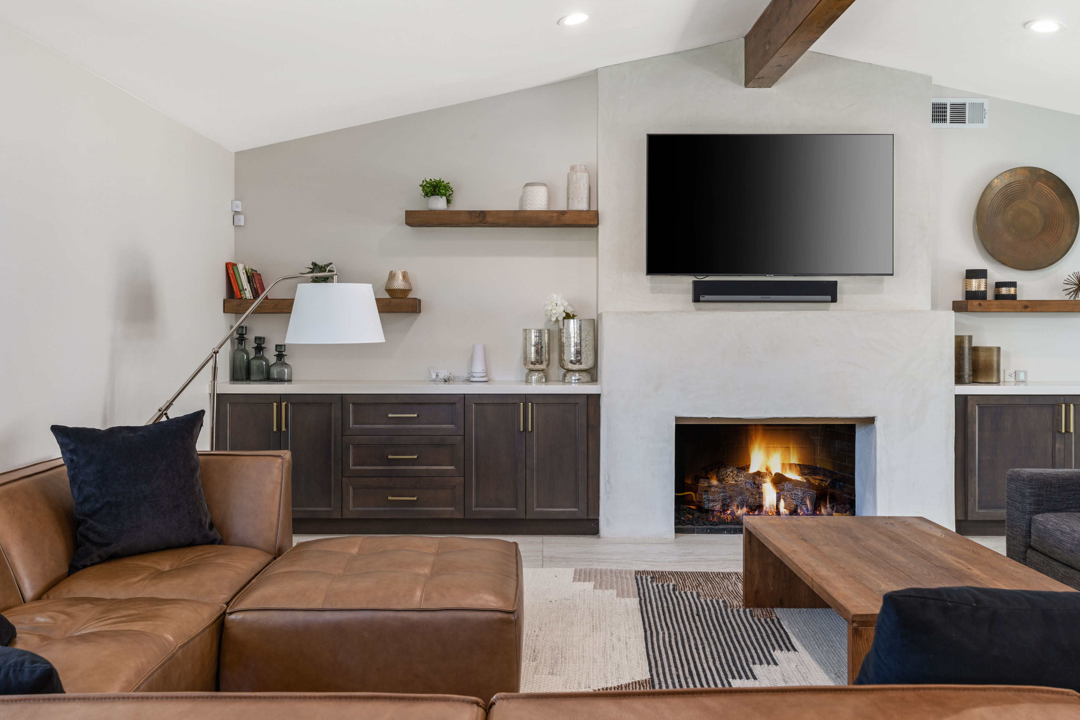 This is a stylish living room featuring a modern fireplace with a wall-mounted TV above it. Brown wooden cabinetry flanks both sides of the fireplace, complemented by sleek countertops and decorative shelves. A sectional couch and a wooden coffee table add to the comfortable and sophisticated atmosphere of the living space.